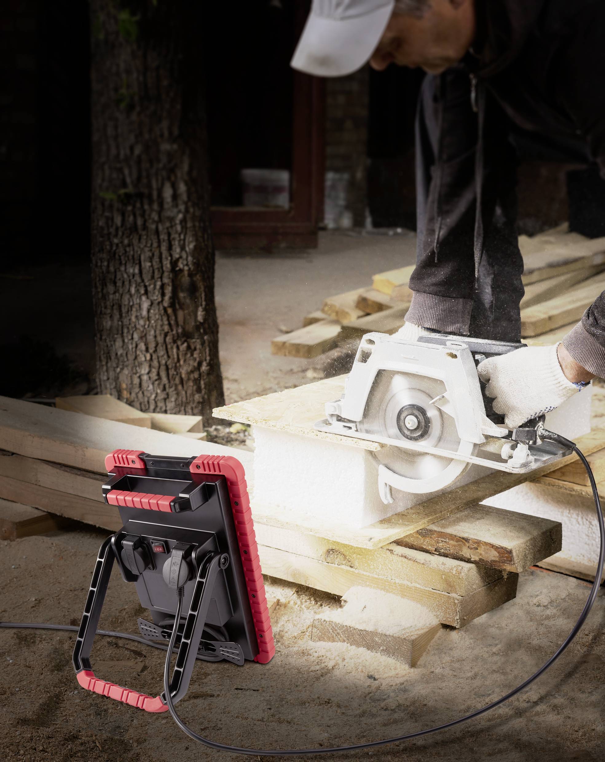 A man is cutting wood with a circular saw. In the foreground, a construction floodlight illuminates the work area.