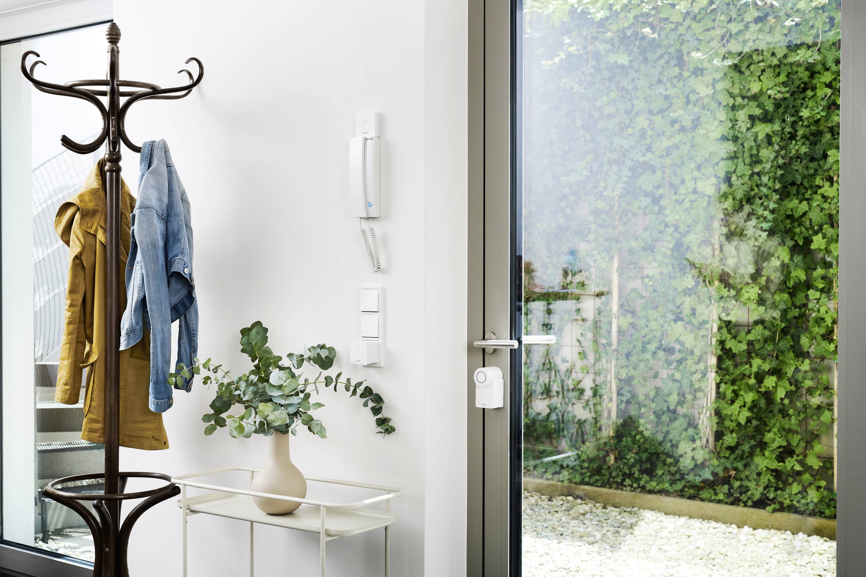 A bright room with a coat stand, on which a yellow jacket and a blue shirt are hanging. Next to it, a table with a vase and plants. A glass door leads to a verdant outdoor area.