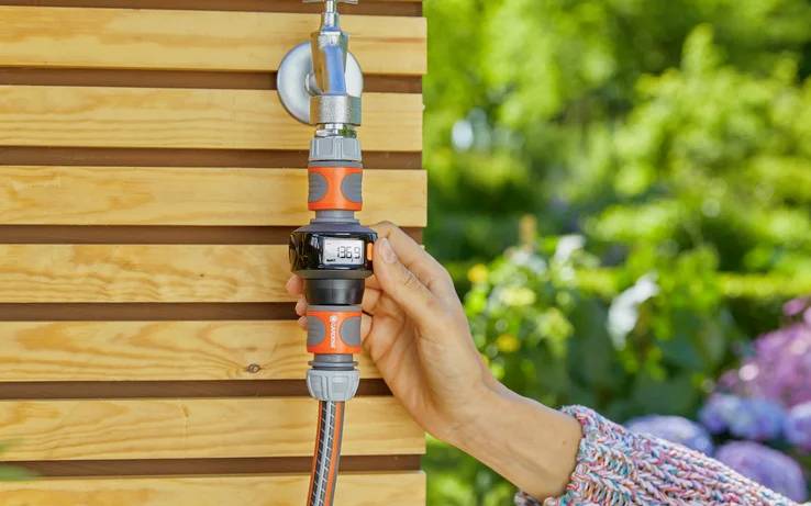 A hand adjusts a digital water timer connected to a hose on a wooden wall outdoors, surrounded by greenery and flowers.