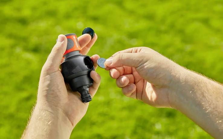 A person inserts a coin into a water flow controller held in their hand against a grassy background.