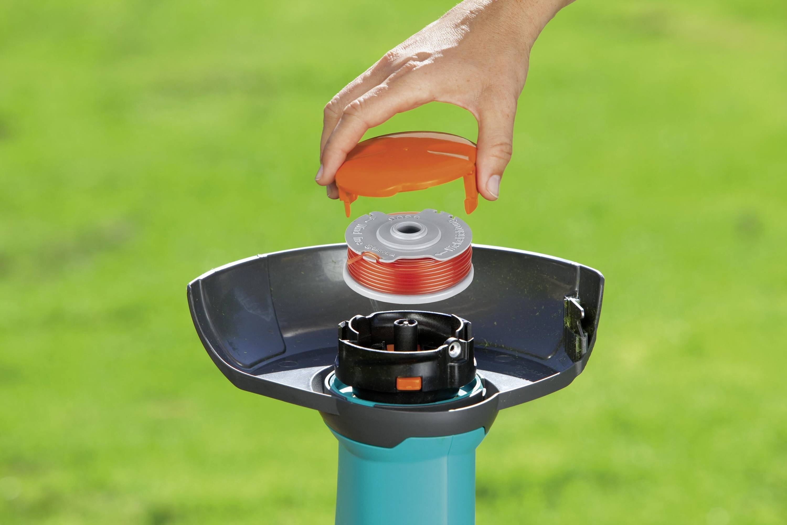 A person is changing the spool of a lawnmower. The lid with the spool is shown. The background is grass green.