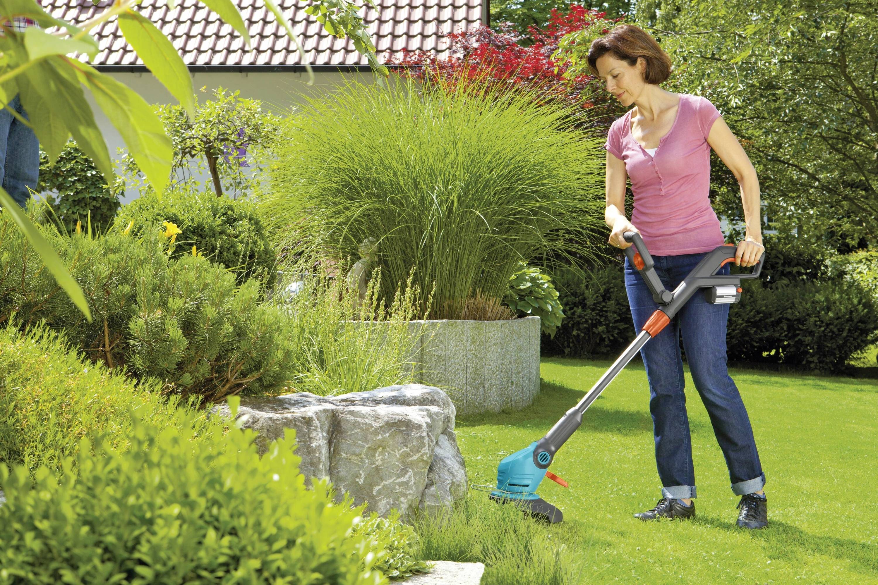 A woman is trimming the lawn in a well-maintained garden with an electric lawn trimmer. Shrubs and a house roof are visible in the background.
