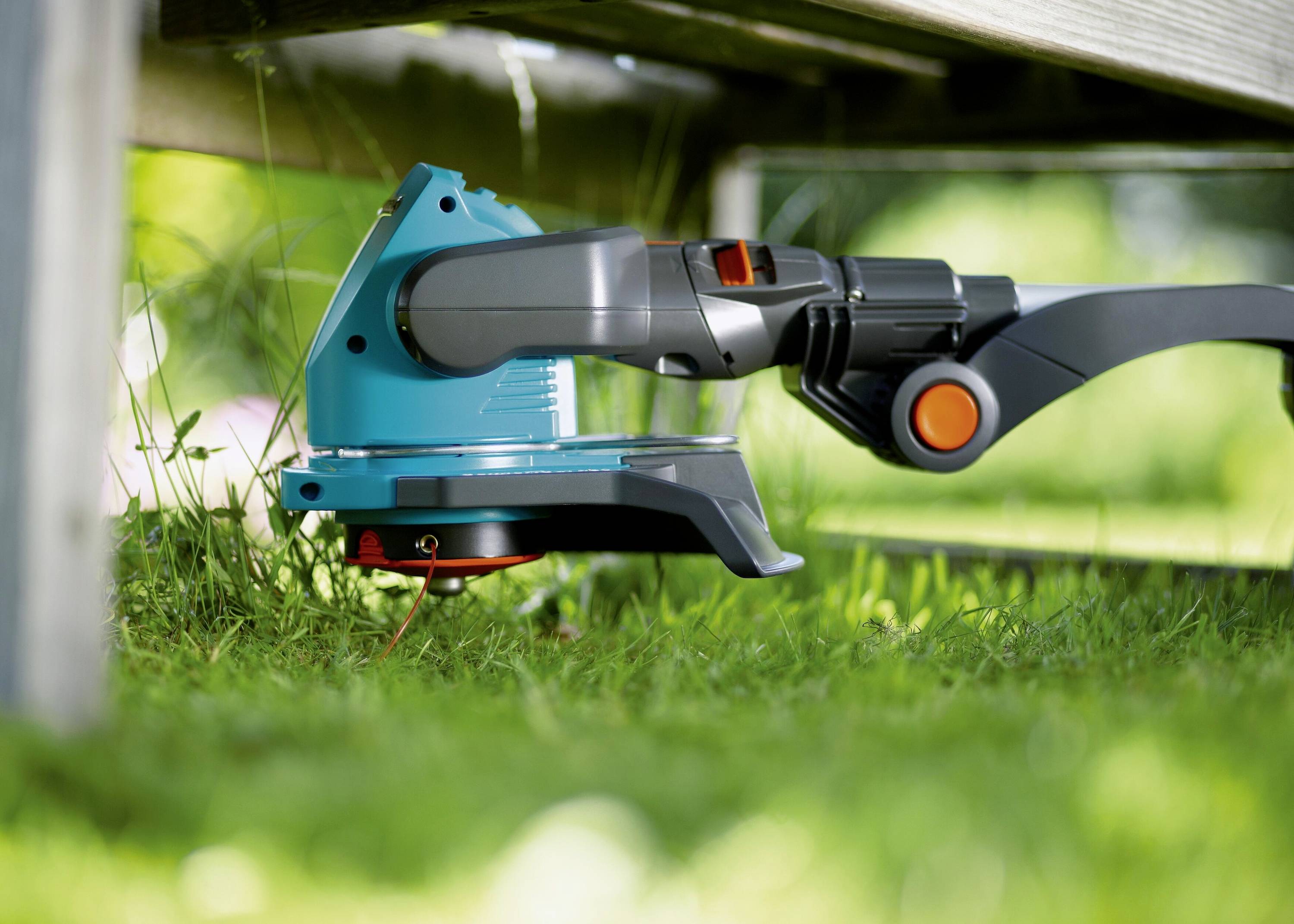An electric lawn trimmer is cutting grass underneath a wooden bench in the garden. The blade is visibly cutting the grass near the edge.
