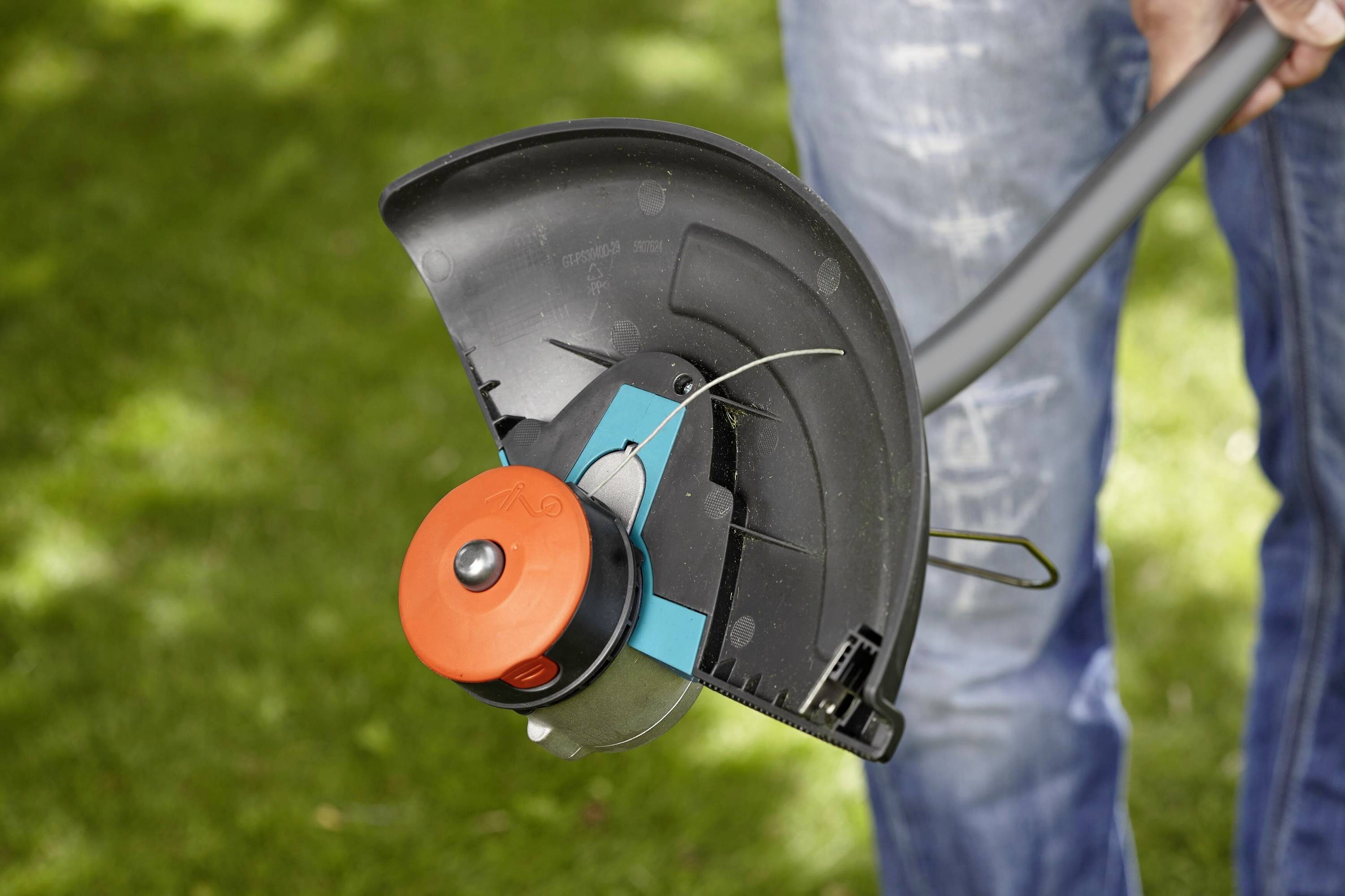 A person is holding a lawn strimmer, with the focus on the cutting head. Grass is blurred in the background.