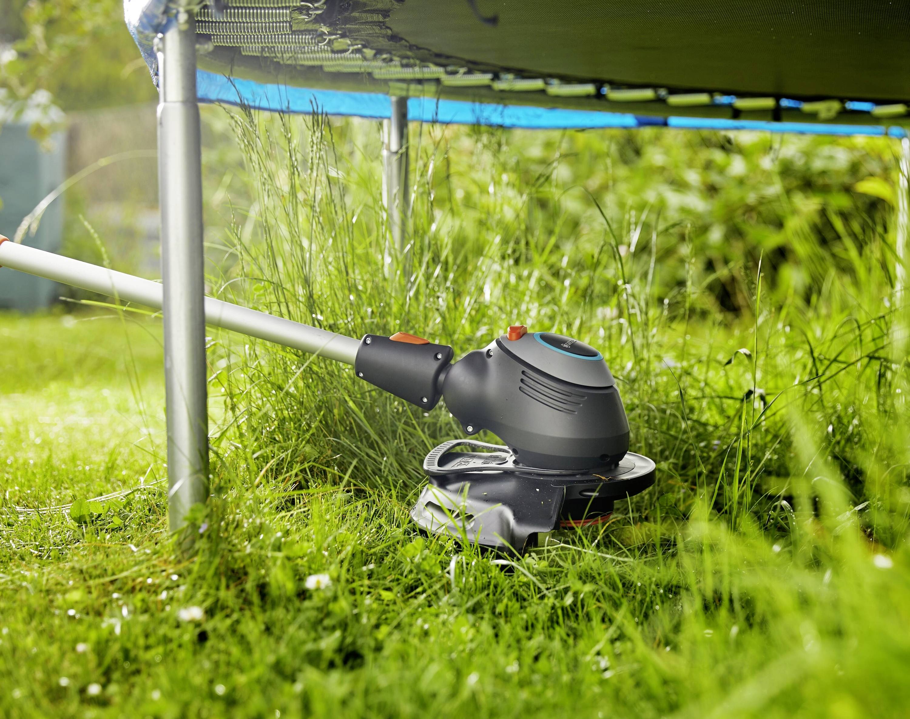 A lawn trimmer is cutting grass underneath a trampoline in the garden. The focus is on precise maintenance in hard-to-reach areas.