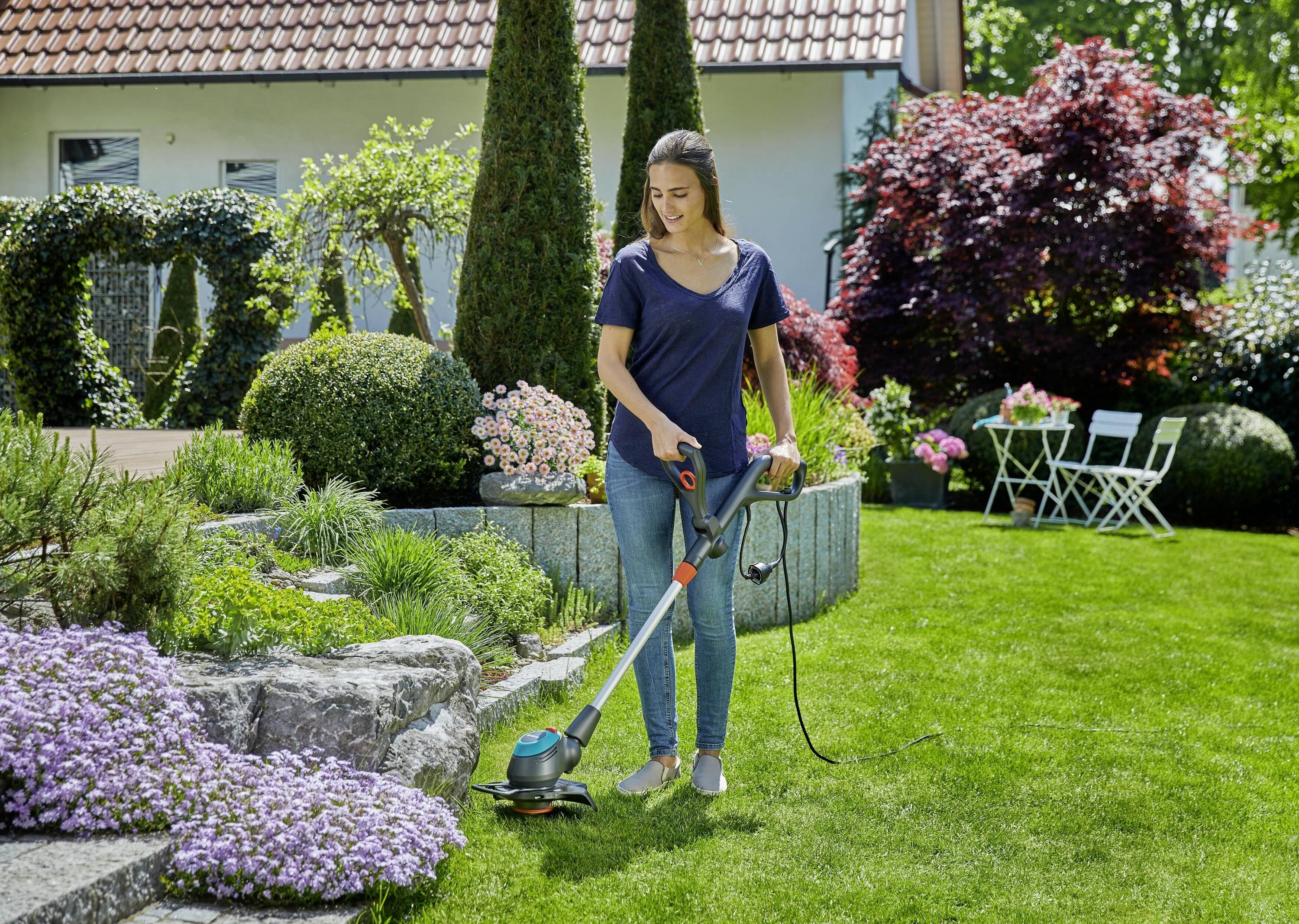 A woman is using a strimmer to trim the grass in a well-maintained garden with flowers and shrubs. A house and garden furniture can be seen in the background.