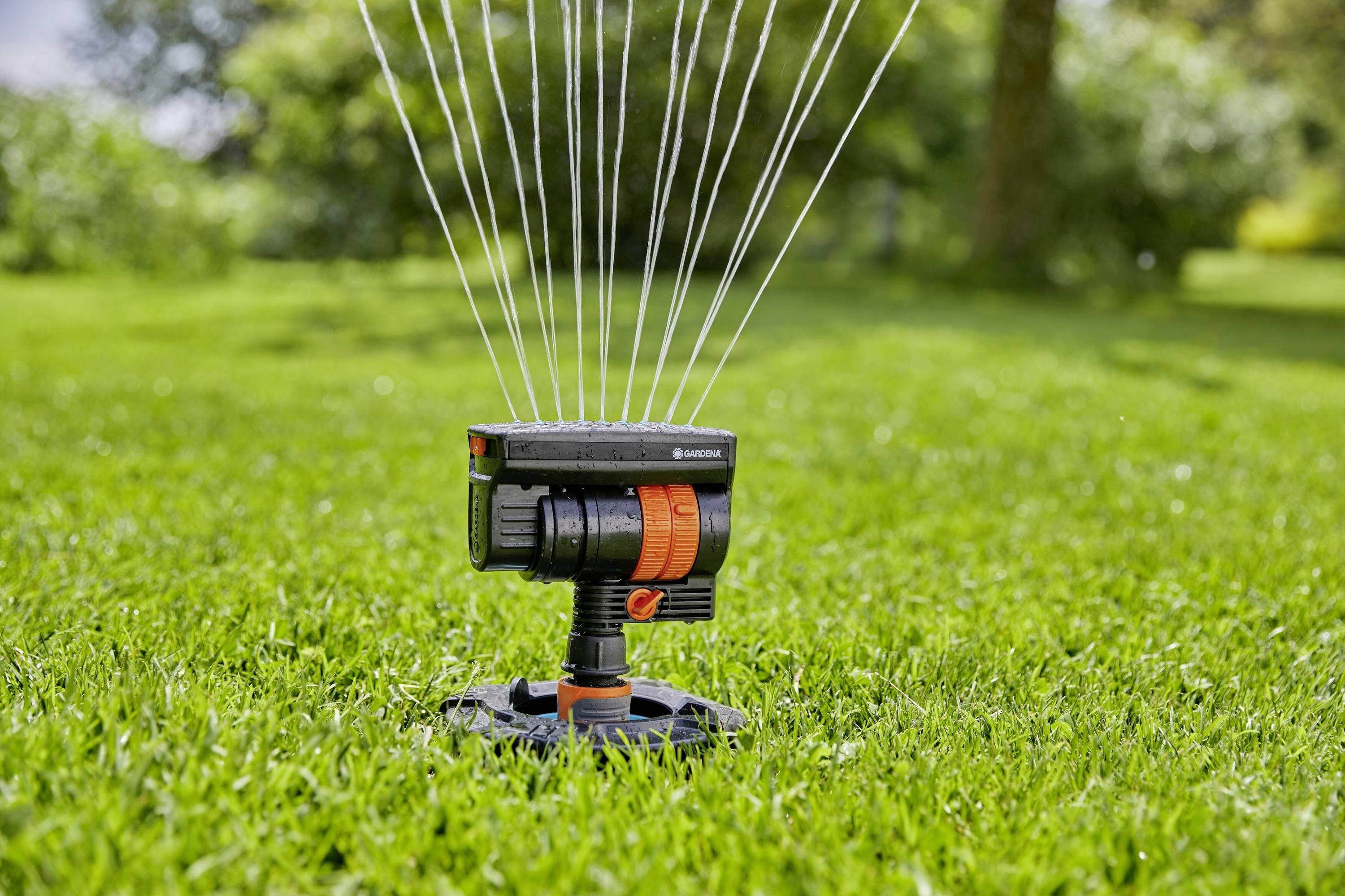 A lawn sprinkler on green grass, projecting multiple water jets into the air to irrigate the surrounding area. Trees are visible in the background.