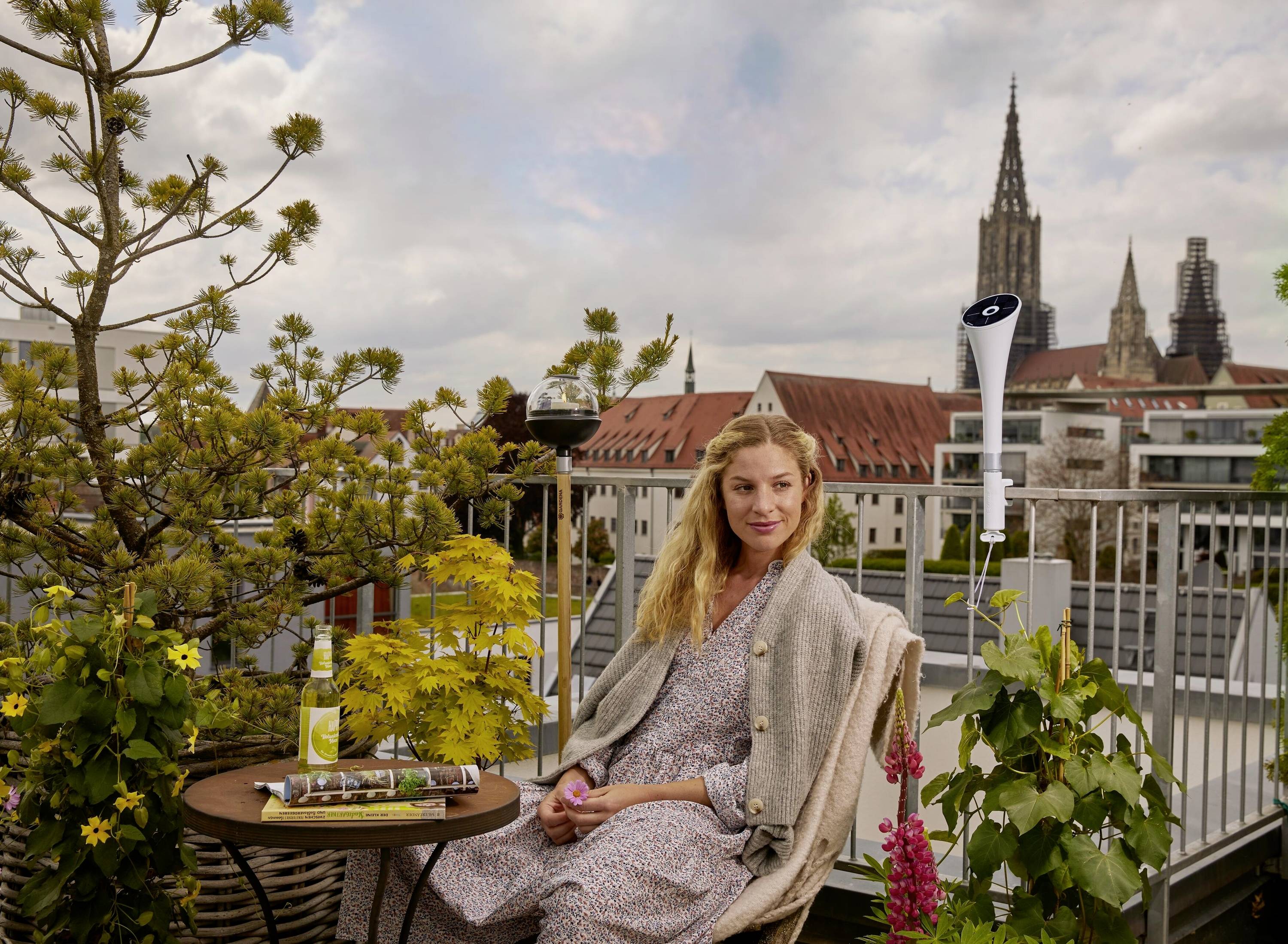 A woman is sitting on a roof terrace with plants and a table, with a city landscape featuring a church spire visible in the background.