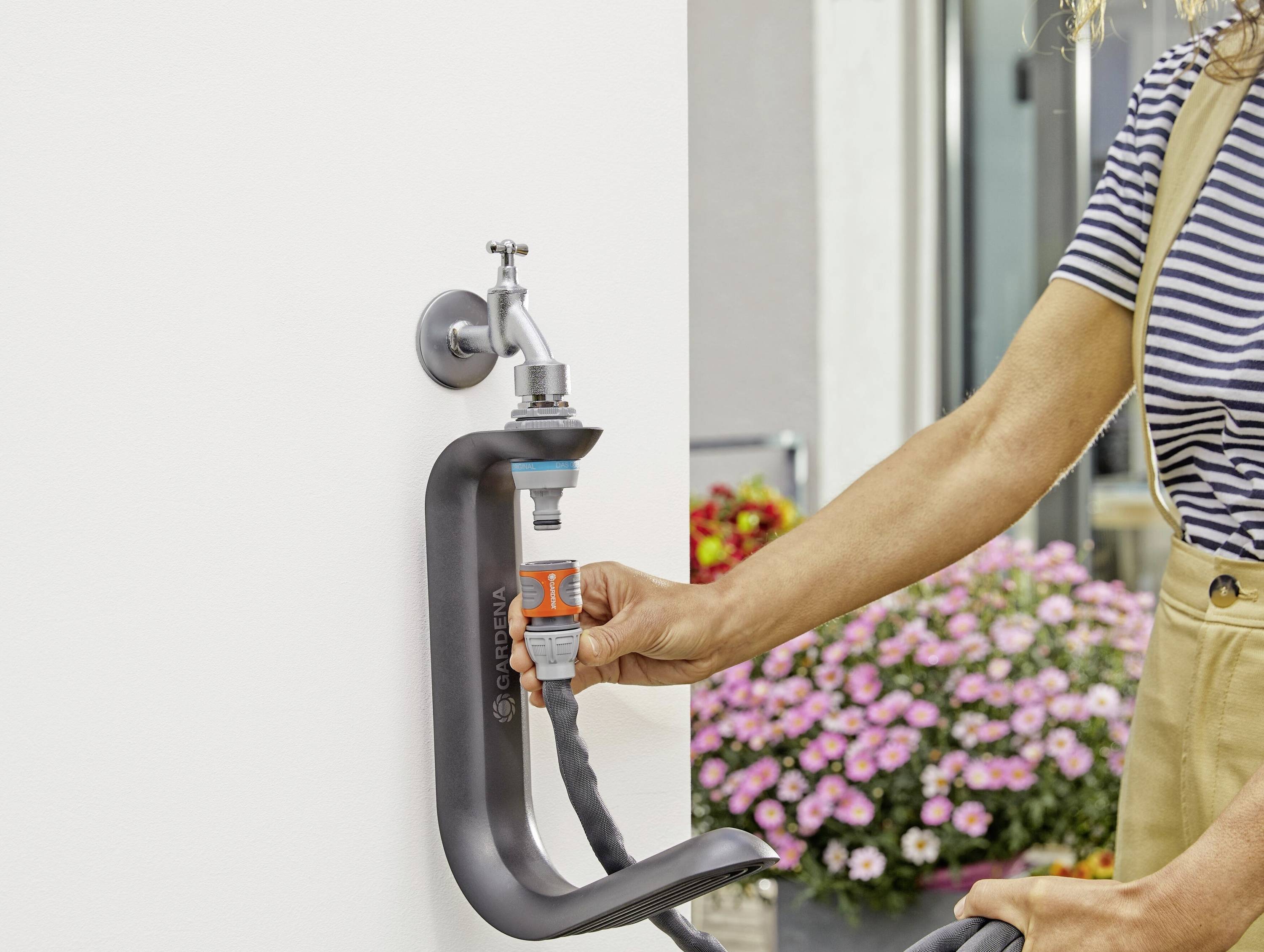 A person connects a garden hose to an outdoor tap. Pink flowers are blooming in a planter in the background.
