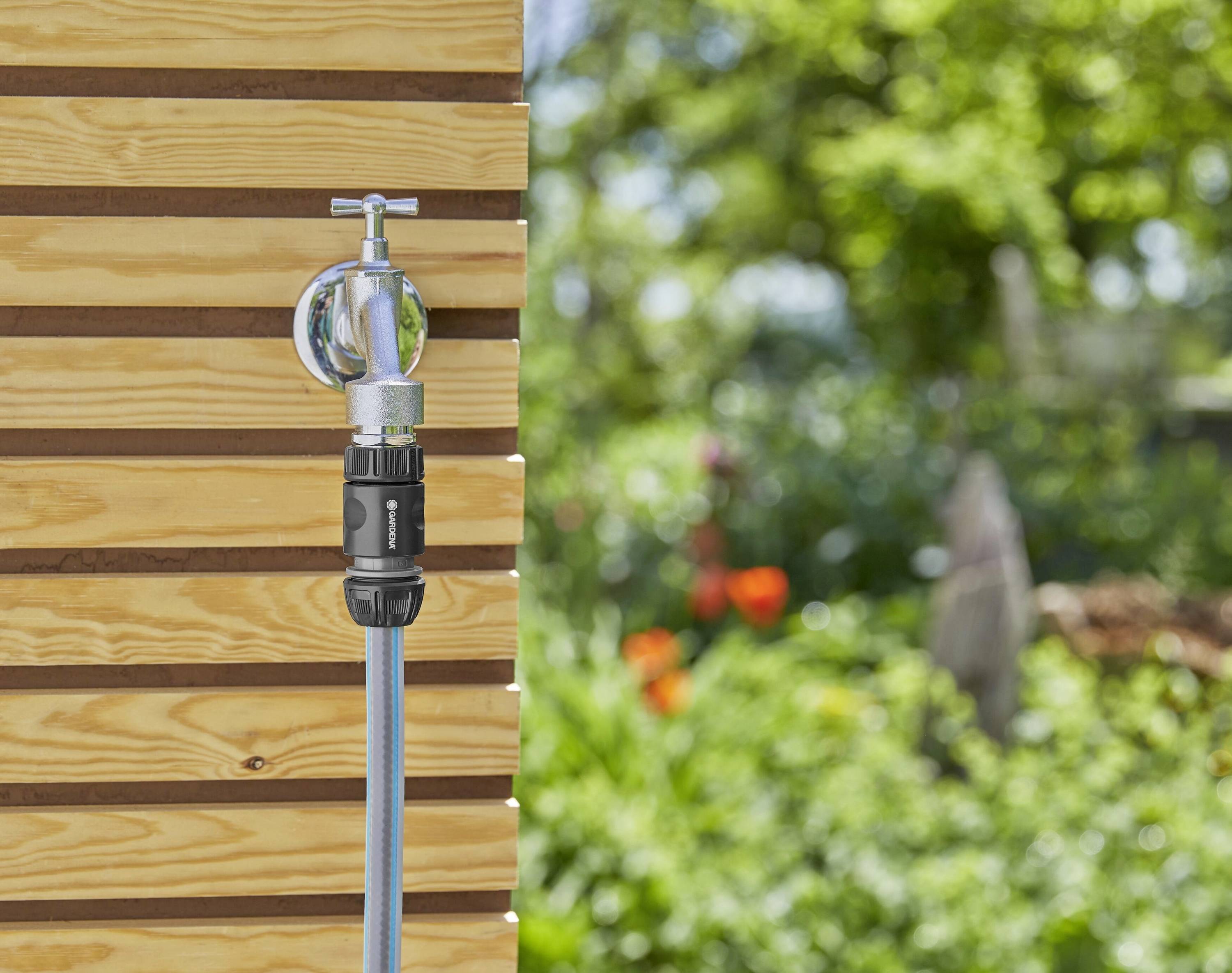 'Garden hose attached to a tap on a wooden wall, surrounded by a green garden with colourful flowers in the background.'