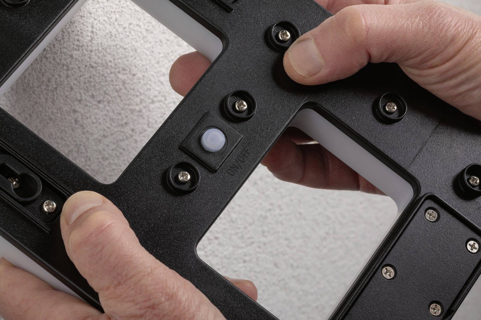 Hands are holding a black plastic part with multiple screws and a central round component.