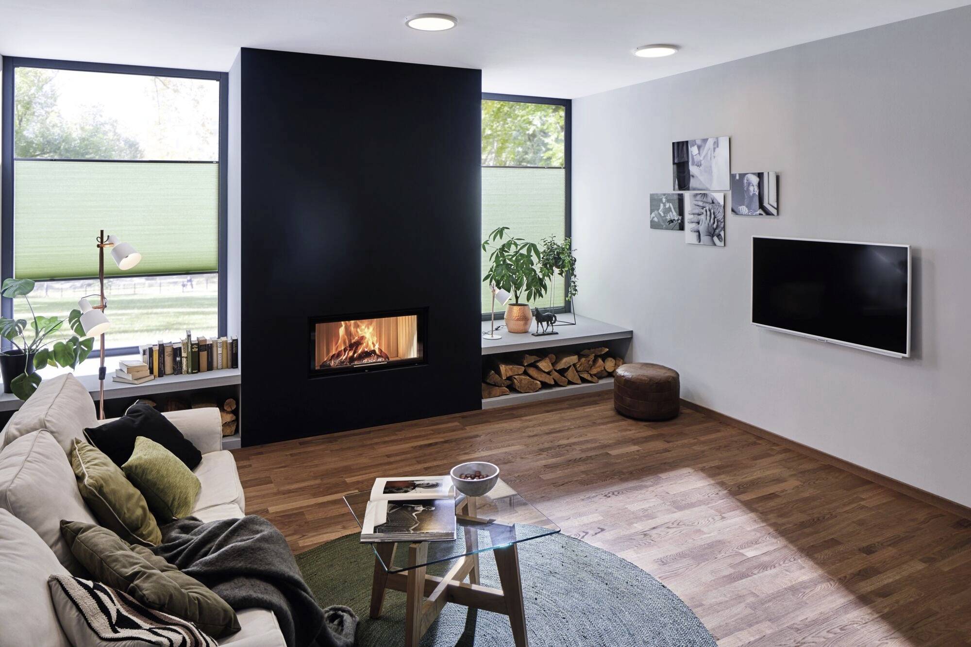 Open-plan living area with a fireplace in a modern design. Cosy seating area, bookshelf, plants and a flatscreen television mounted on the wall.