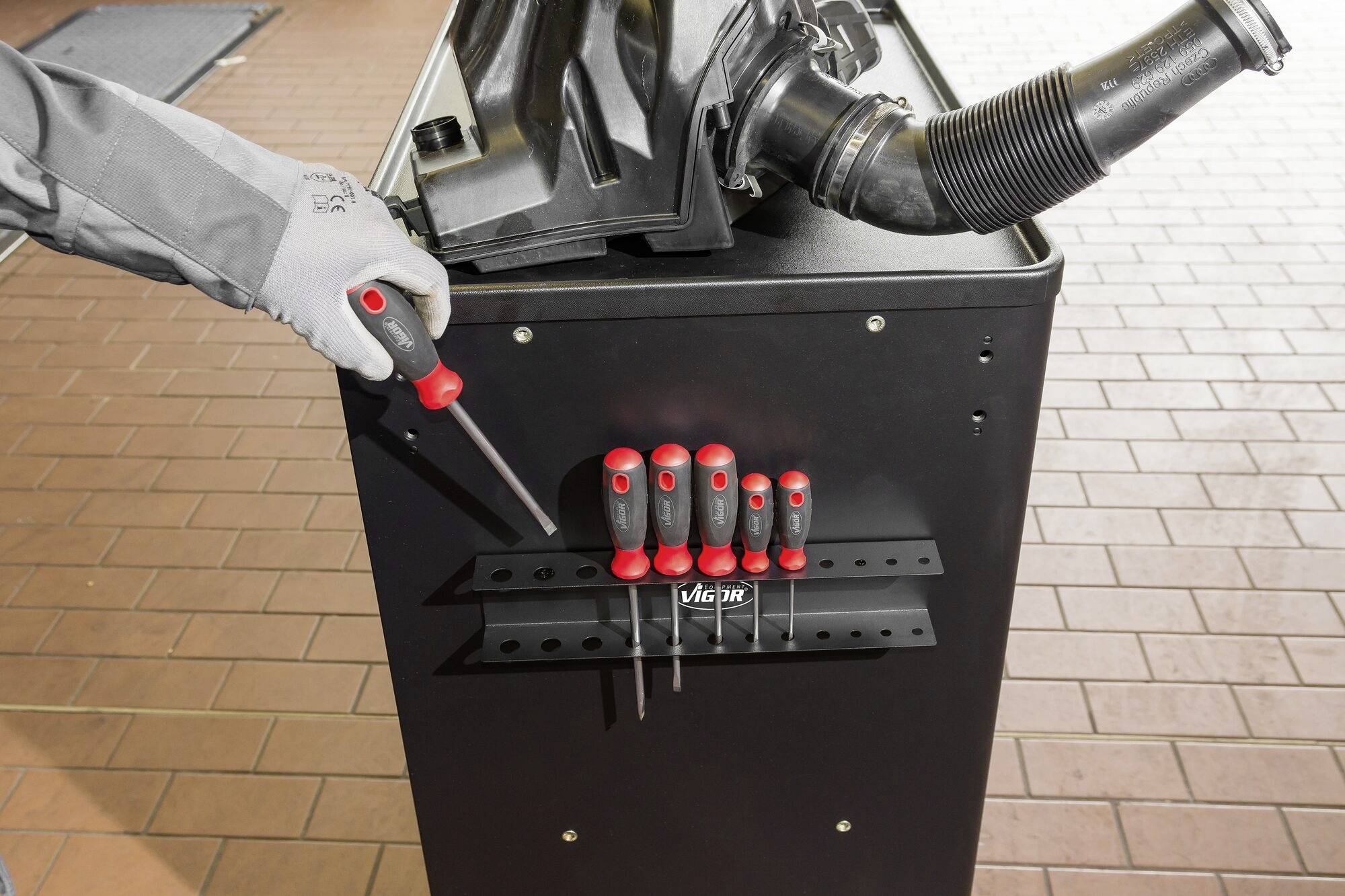 A hand in a grey glove reaches for a screwdriver from a tool trolley with multiple screwdrivers in front of a workbench.