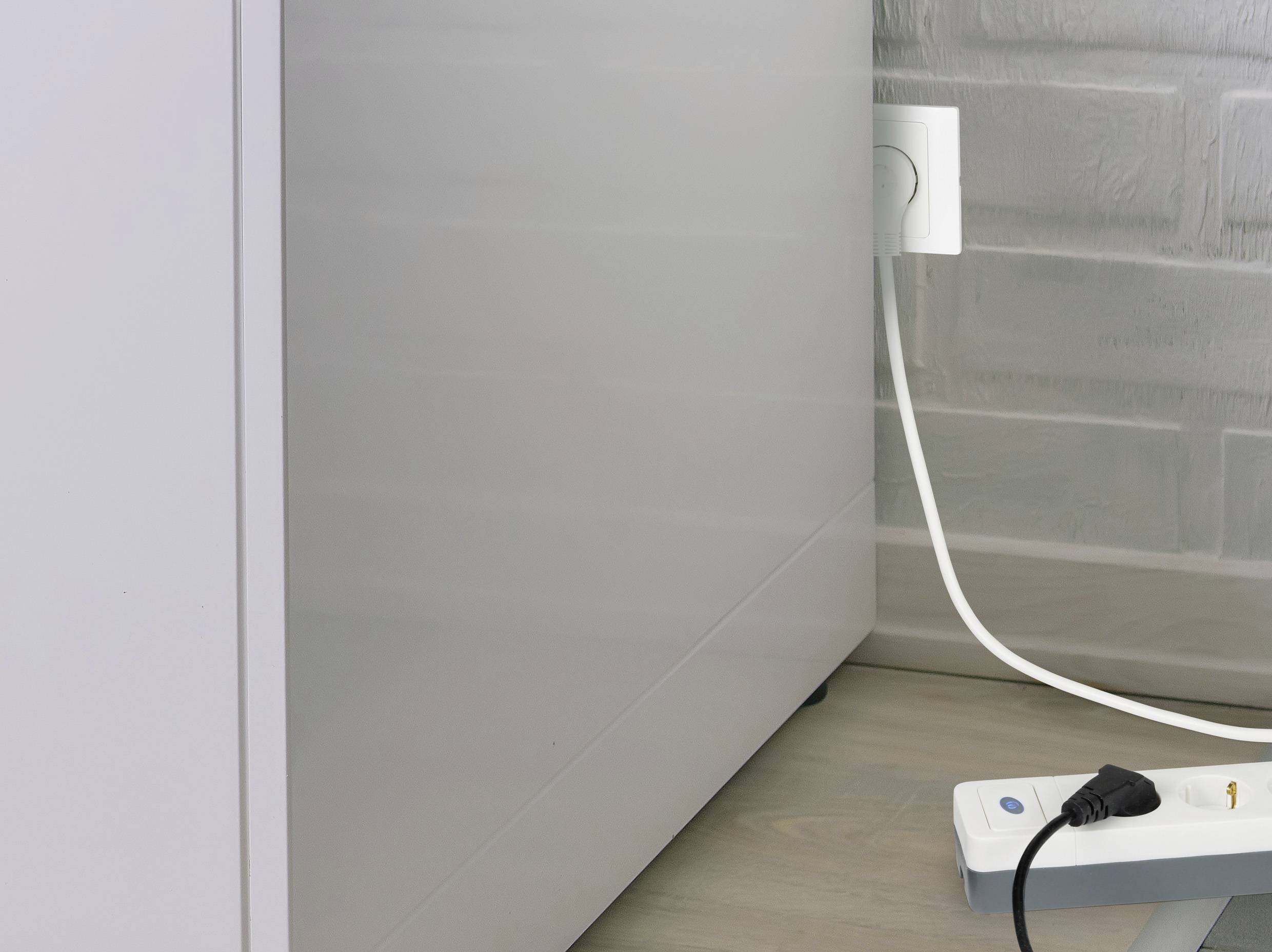 A white cable is plugged into a wall socket, with a grey power strip lying on the floor in front of a grey wall.