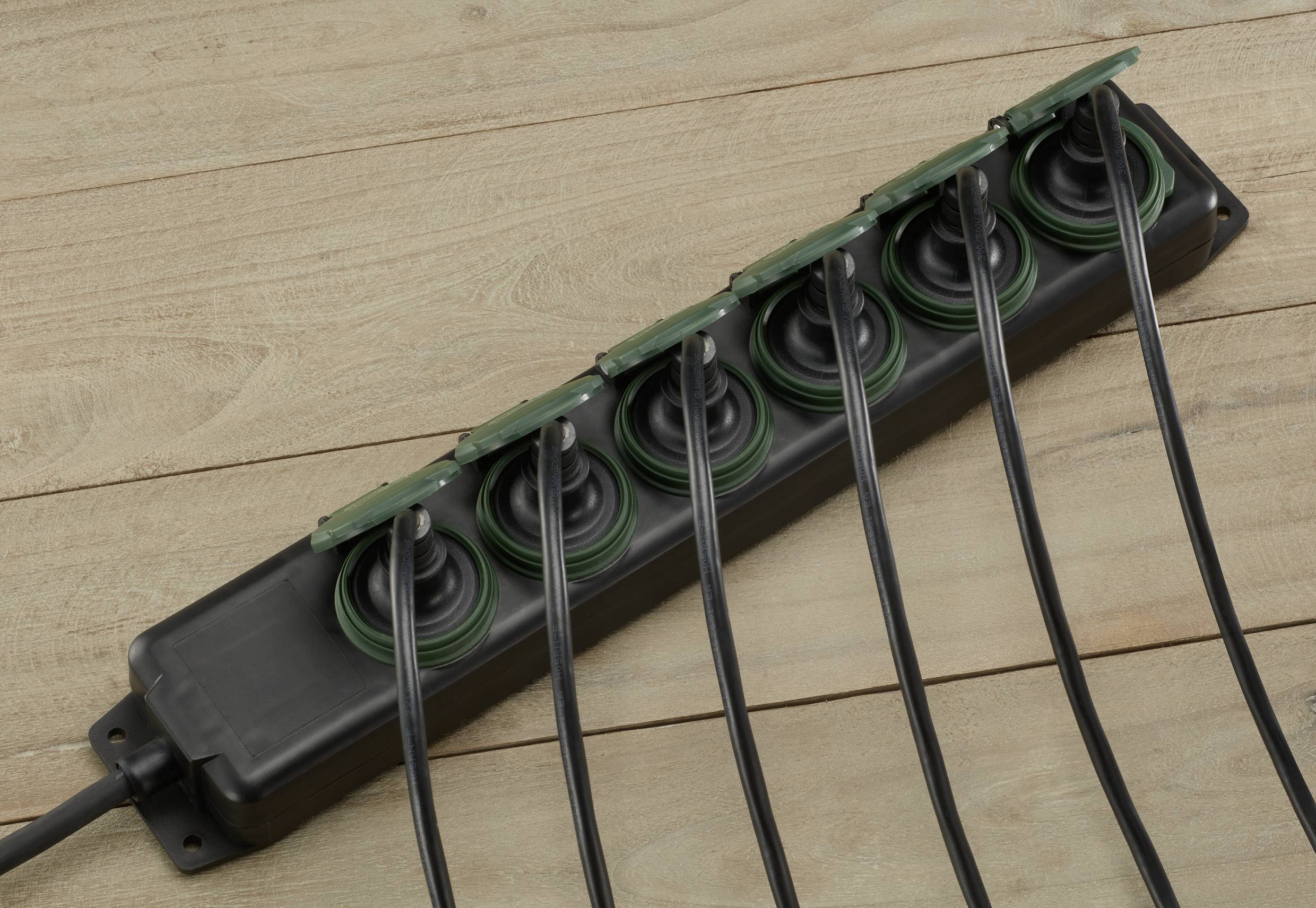 Multi-socket extension lead in black plastic with six socket outlets and green circles, lying on a wooden floor.