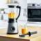 A blender with chopped fruits on a wooden worktop in a modern kitchen. Next to it, a glass of smoothie and a pestle.