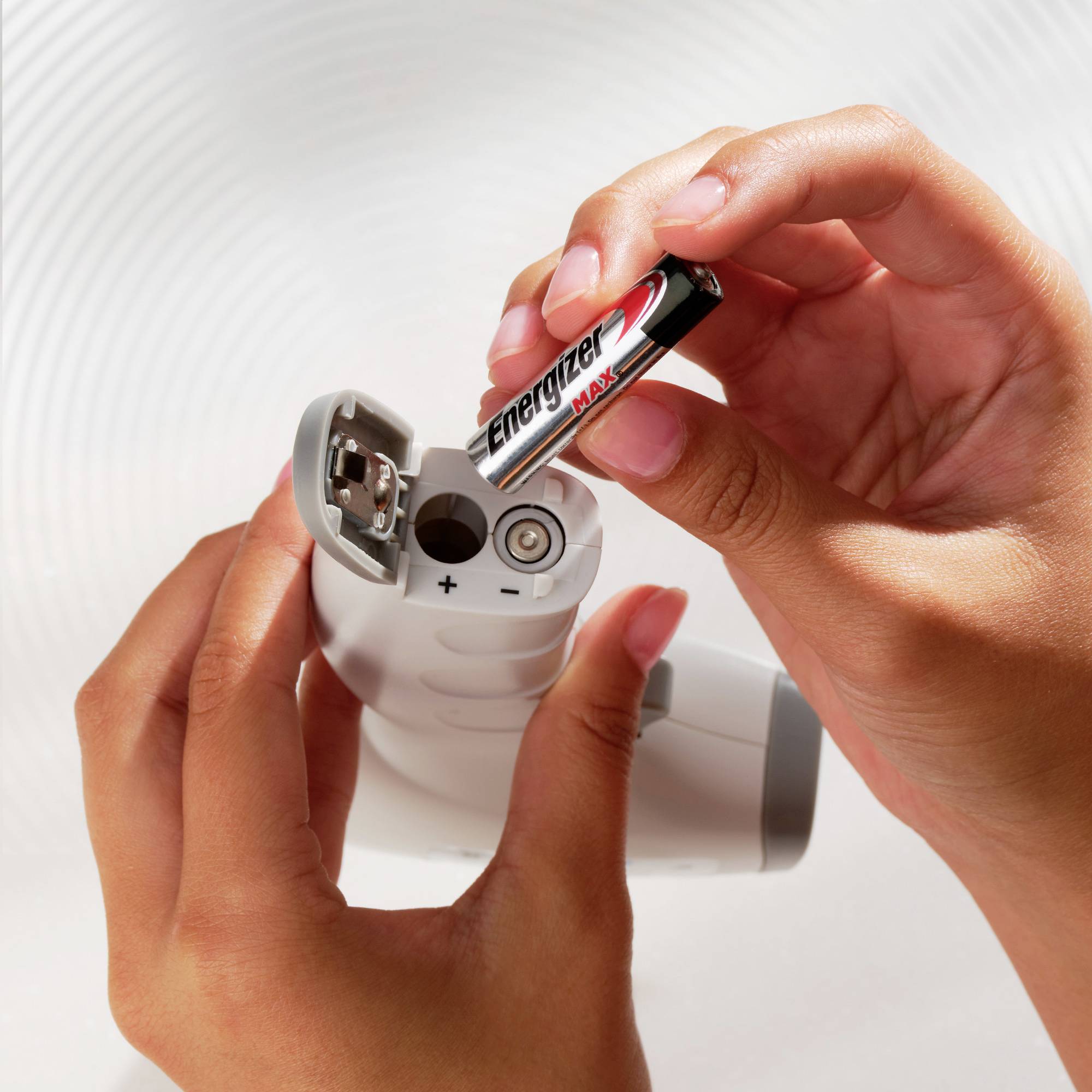 A person is inserting an 'Energizer Max' battery into a device. Hands are holding the battery and the device. The light is bright and focused.