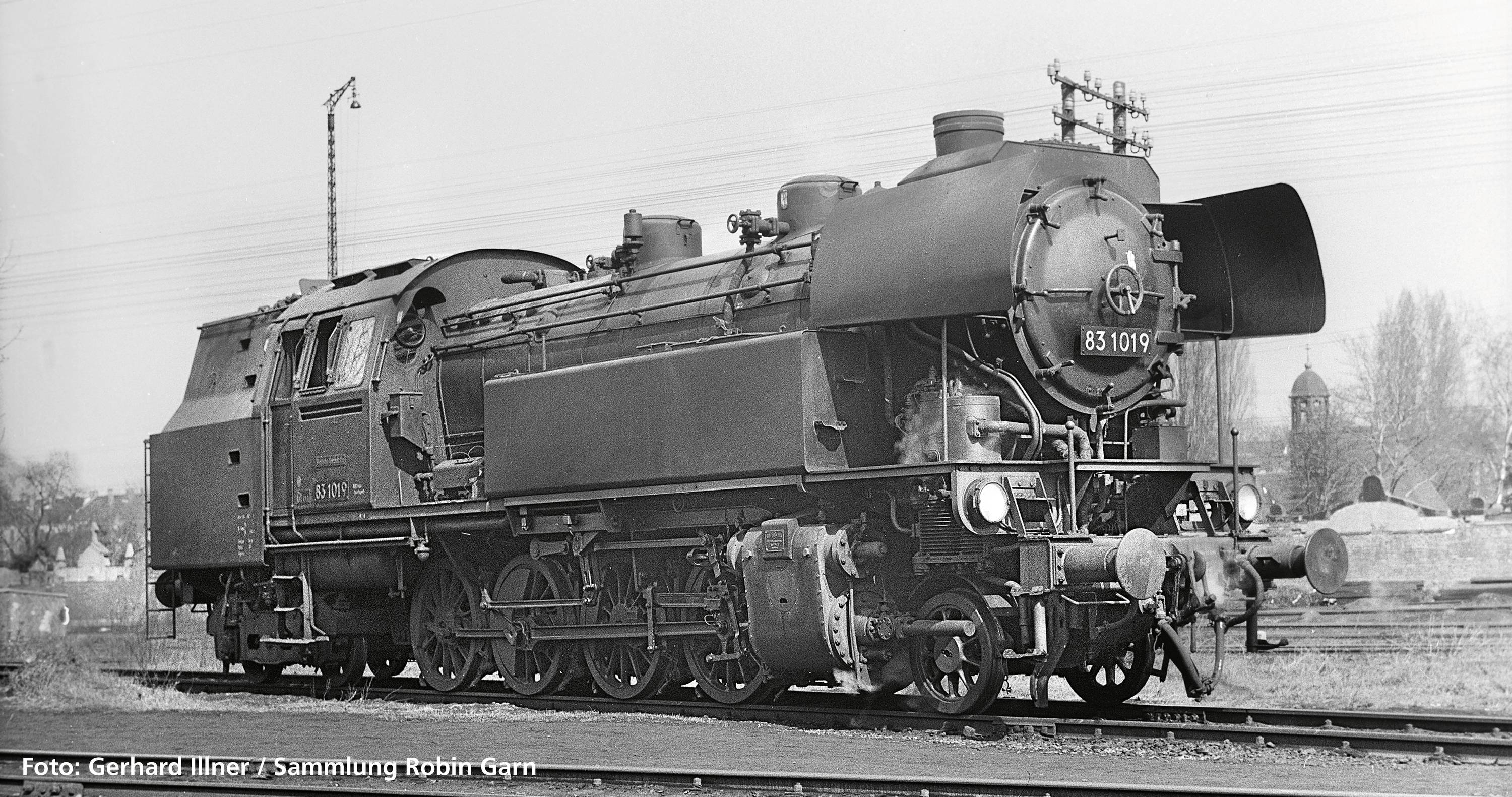 A steam locomotive of class 83.10 stands on a railway track. Trees and building structures are visible in the background.