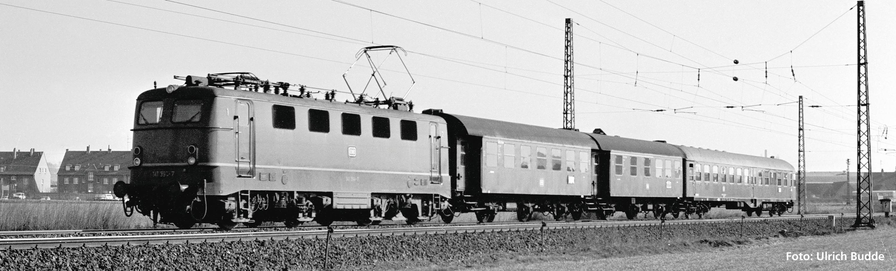 Black and white photograph of a train with a locomotive and two carriages on railway tracks, overhead power lines, with houses in the background.