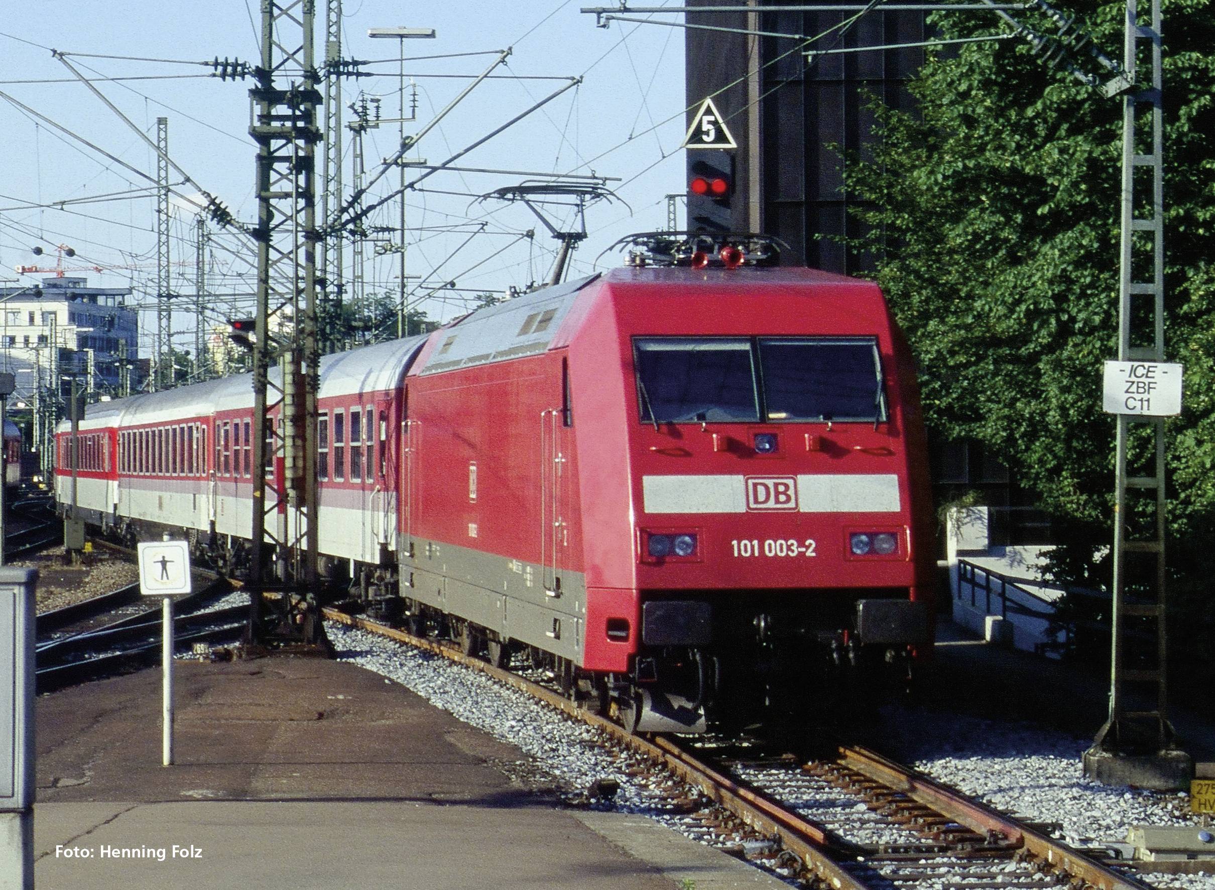 A red Deutsche Bahn train is travelling on a track in an urban setting. Overhead lines and signals are visible on the left side of the image.