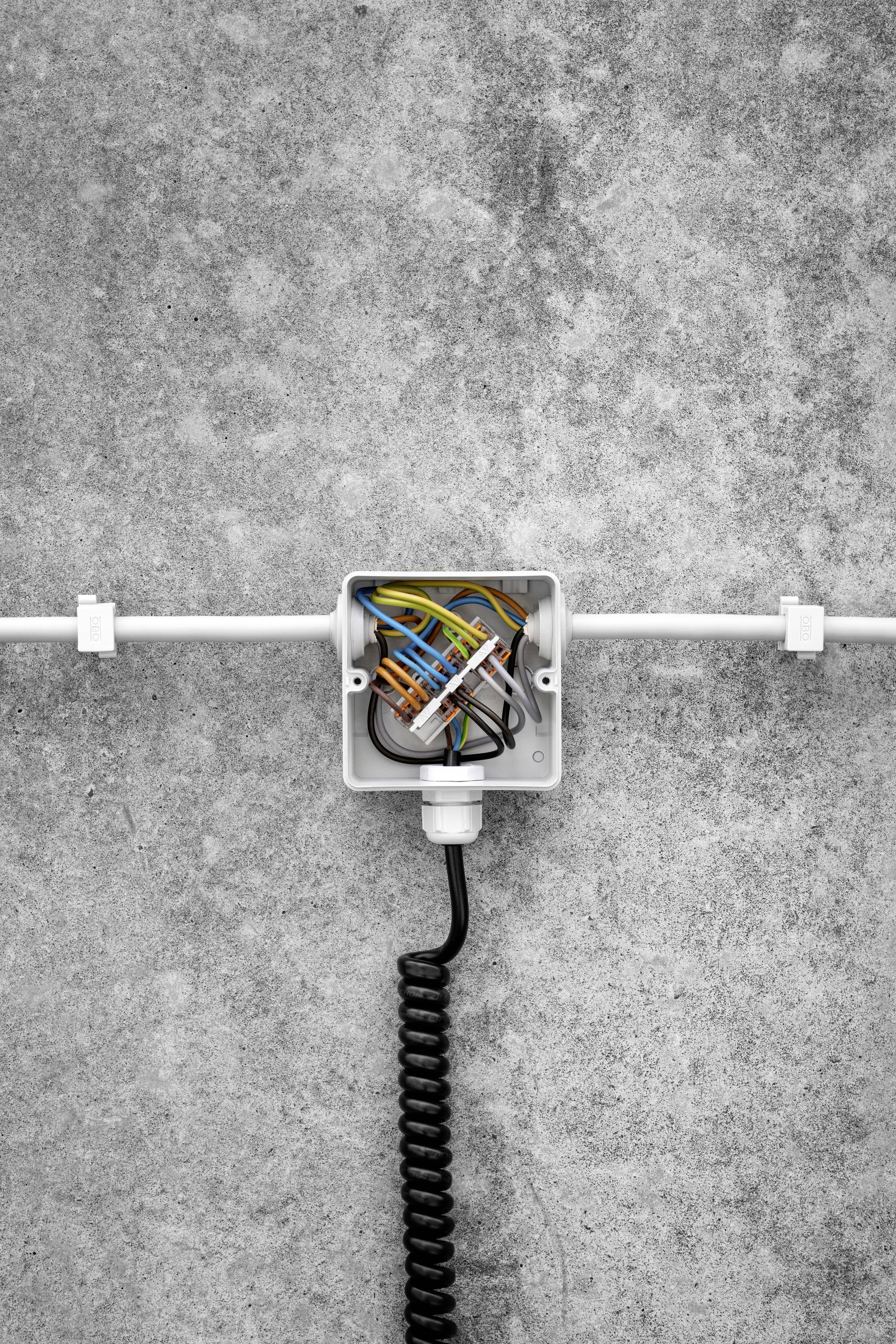 An open distribution box with exposed, coloured wires is mounted on a concrete wall. A black cable runs downwards.