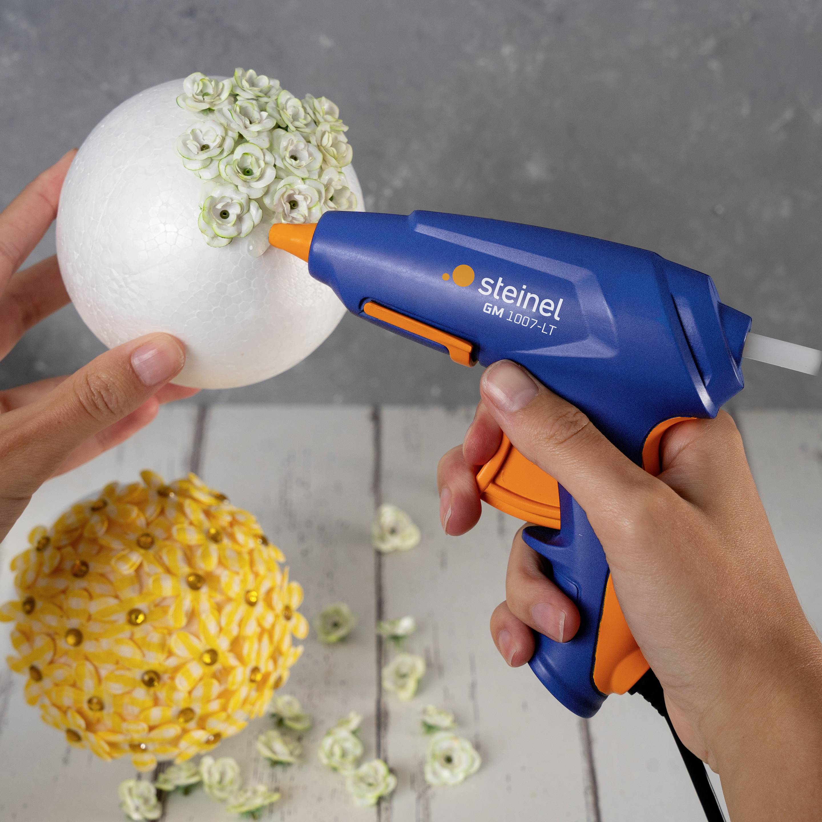 A person is using a hot glue gun to stick small flowers onto a decorative sphere. A finished sphere lies beside it.