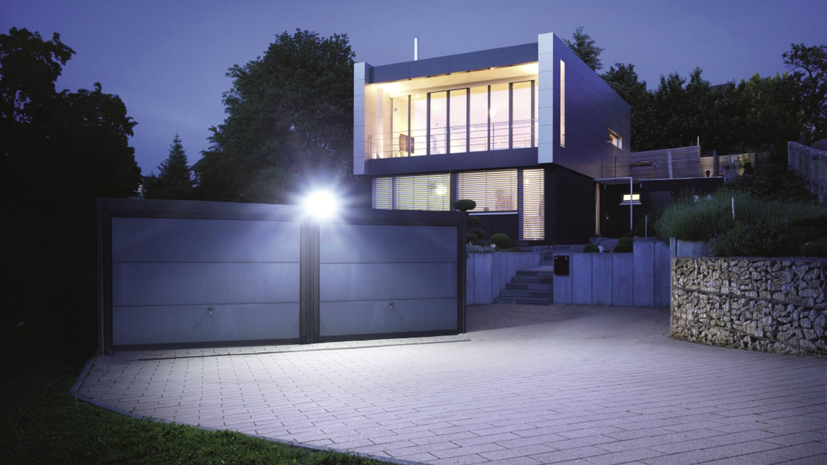 Modern two-storey villa with a large glass façade, illuminated at night. In front of the house, a wide driveway with a closed gate.