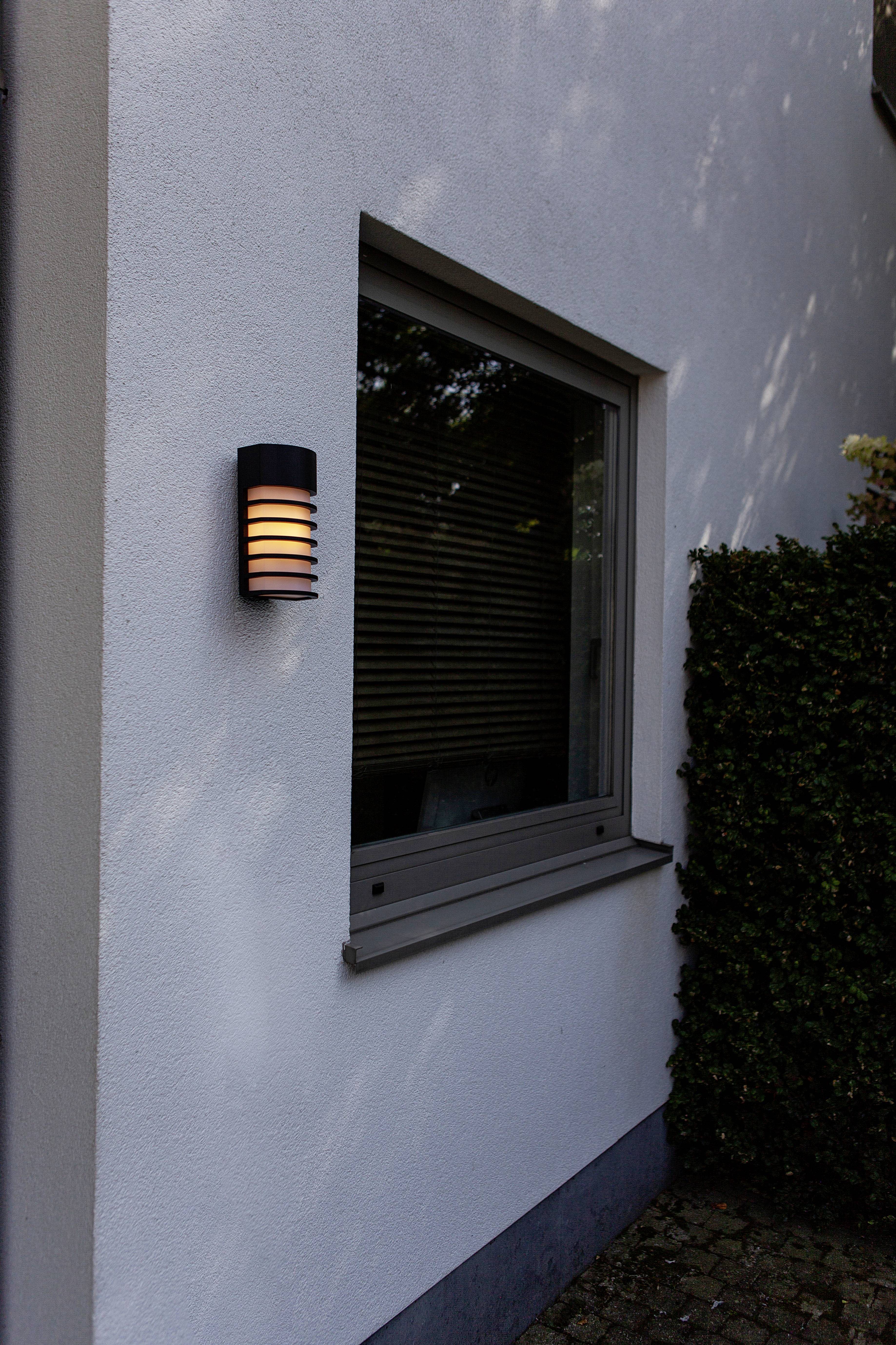 Exterior wall light mounted beside a window on the house facade, with a modern design. Illuminating an area of the pavement in front of a bush.