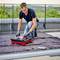 A person is kneeling on a terrace and working intently with a small tiling machine, surrounded by other tools.