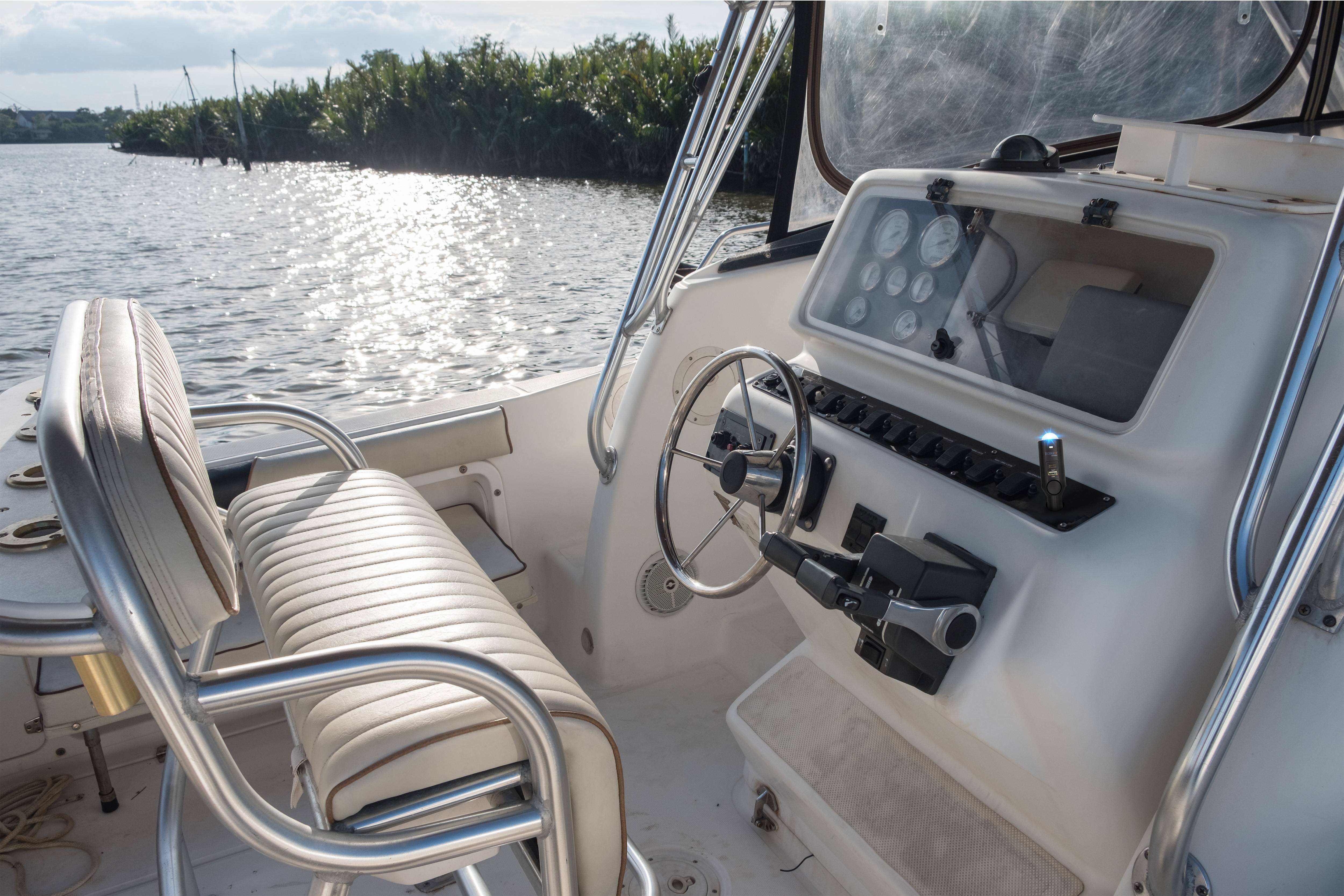 Two padded boat seats in front of a helm with a steering wheel and instruments. View of a river with plants along the bank.