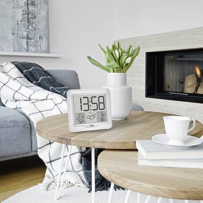 A digital alarm clock shows 13:58 and sits on a table in a cosy living room with a sofa, fireplace, and plant.