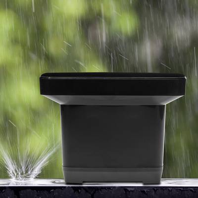 A black plant pot sits outside in the rain. Water droplets bounce off the rim of the pot. The background is blurred.