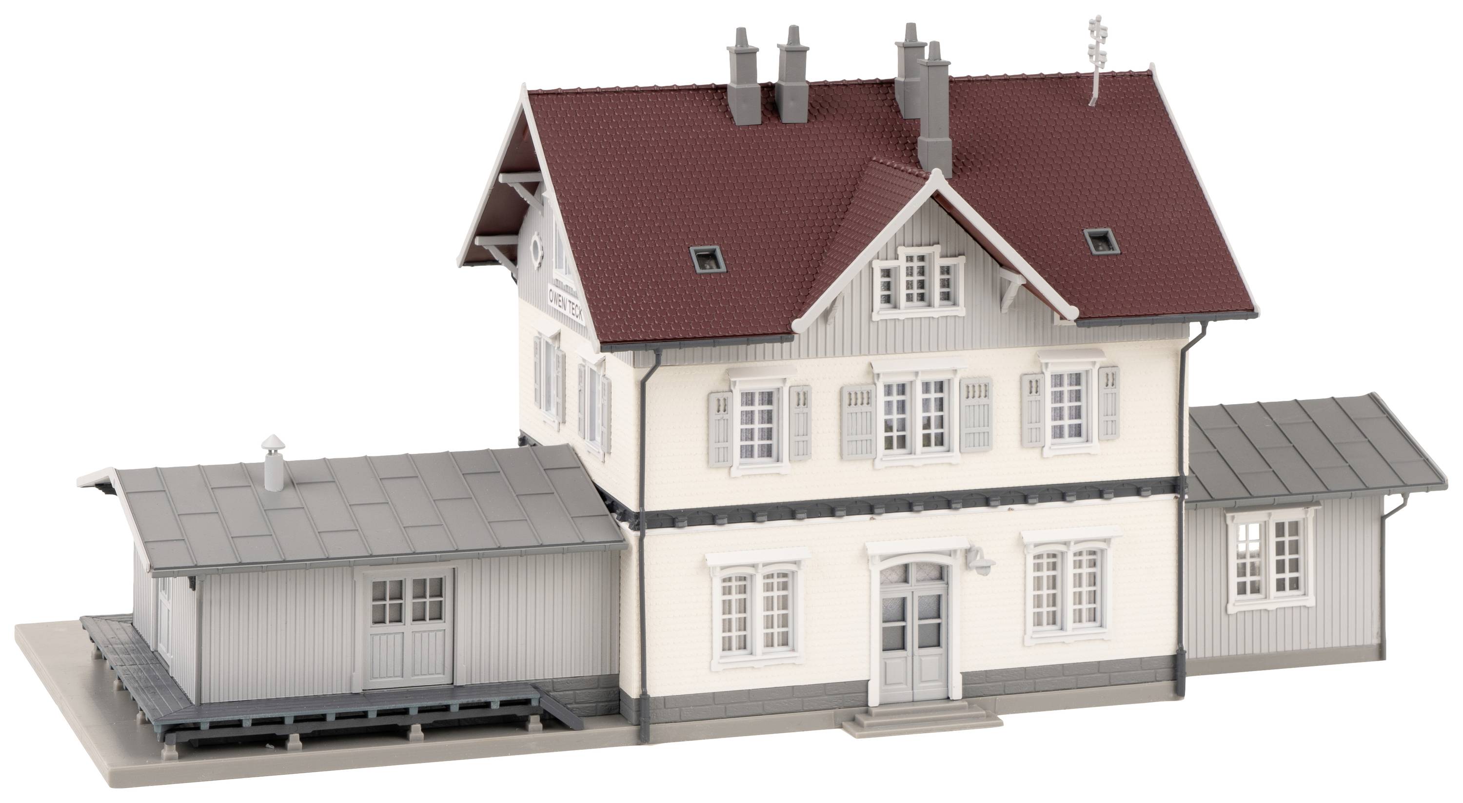 A model building with a red roof and grey extension, possibly a small railway station building, on a display plate.