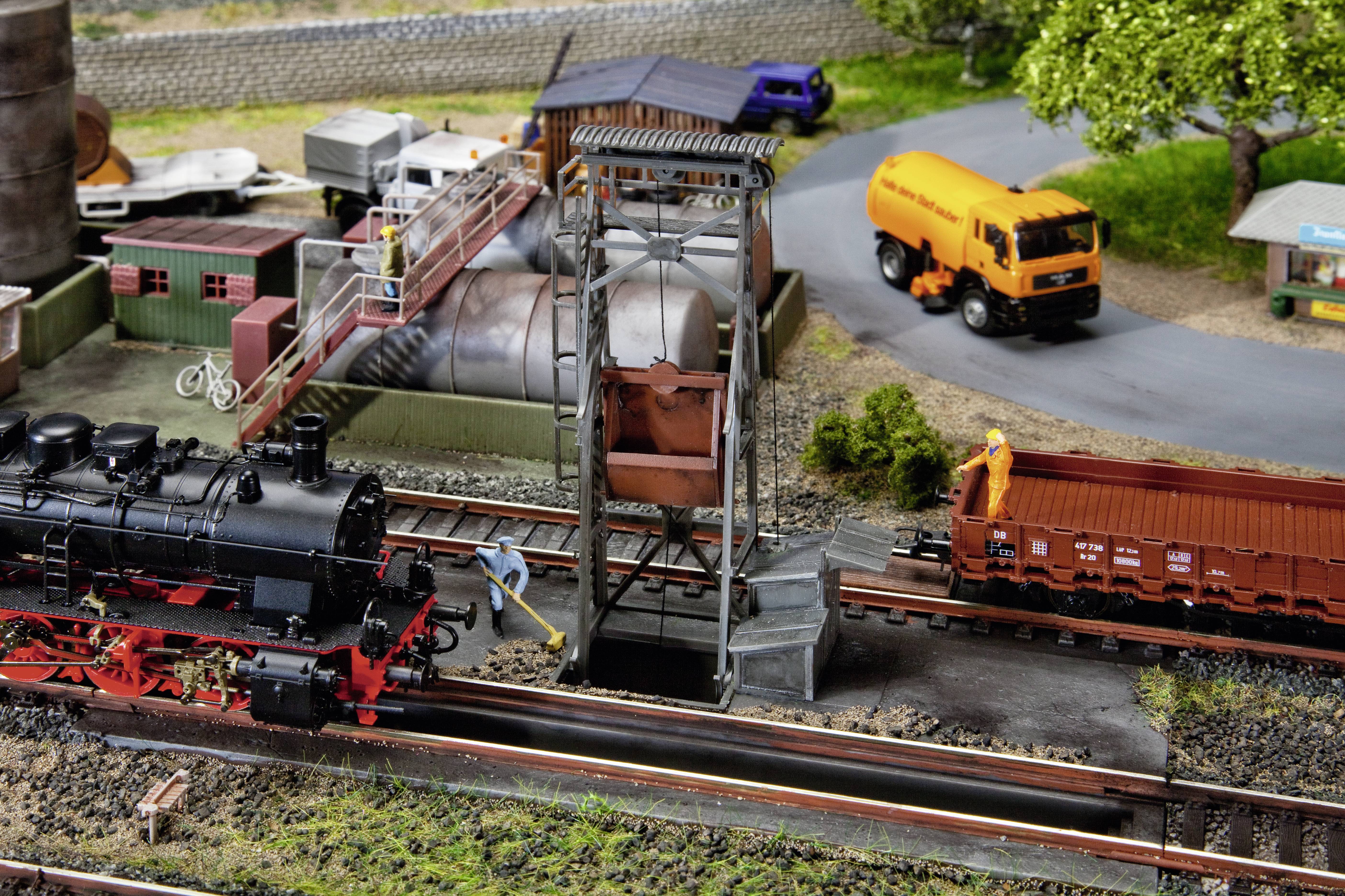 Model railway scene with a steam locomotive stopped next to a crane. A lorry and goods wagons are visible in the background.