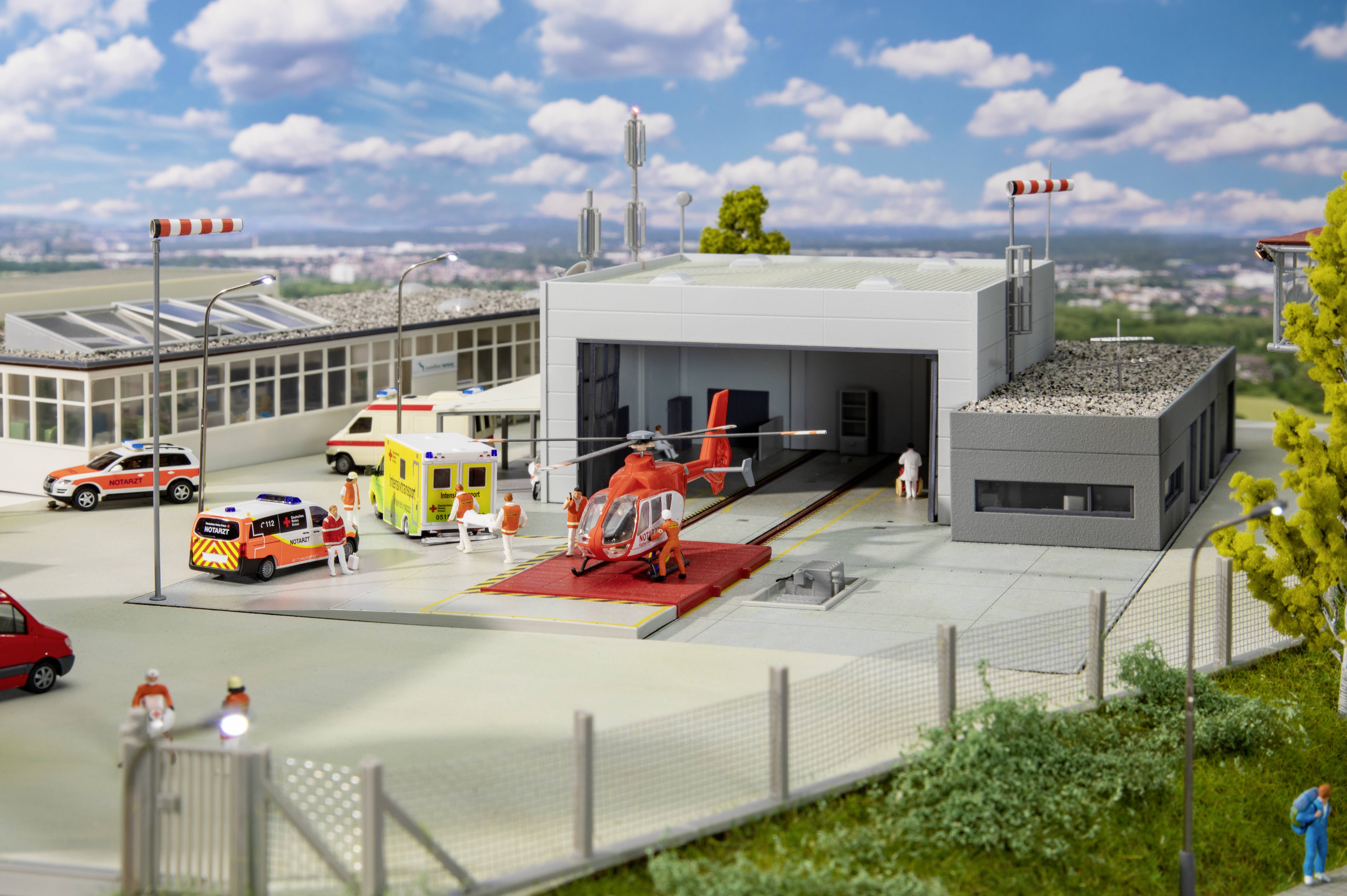 A miniature helicopter on a helipad in front of a hangar. Emergency vehicles and figures are scattered around the building.