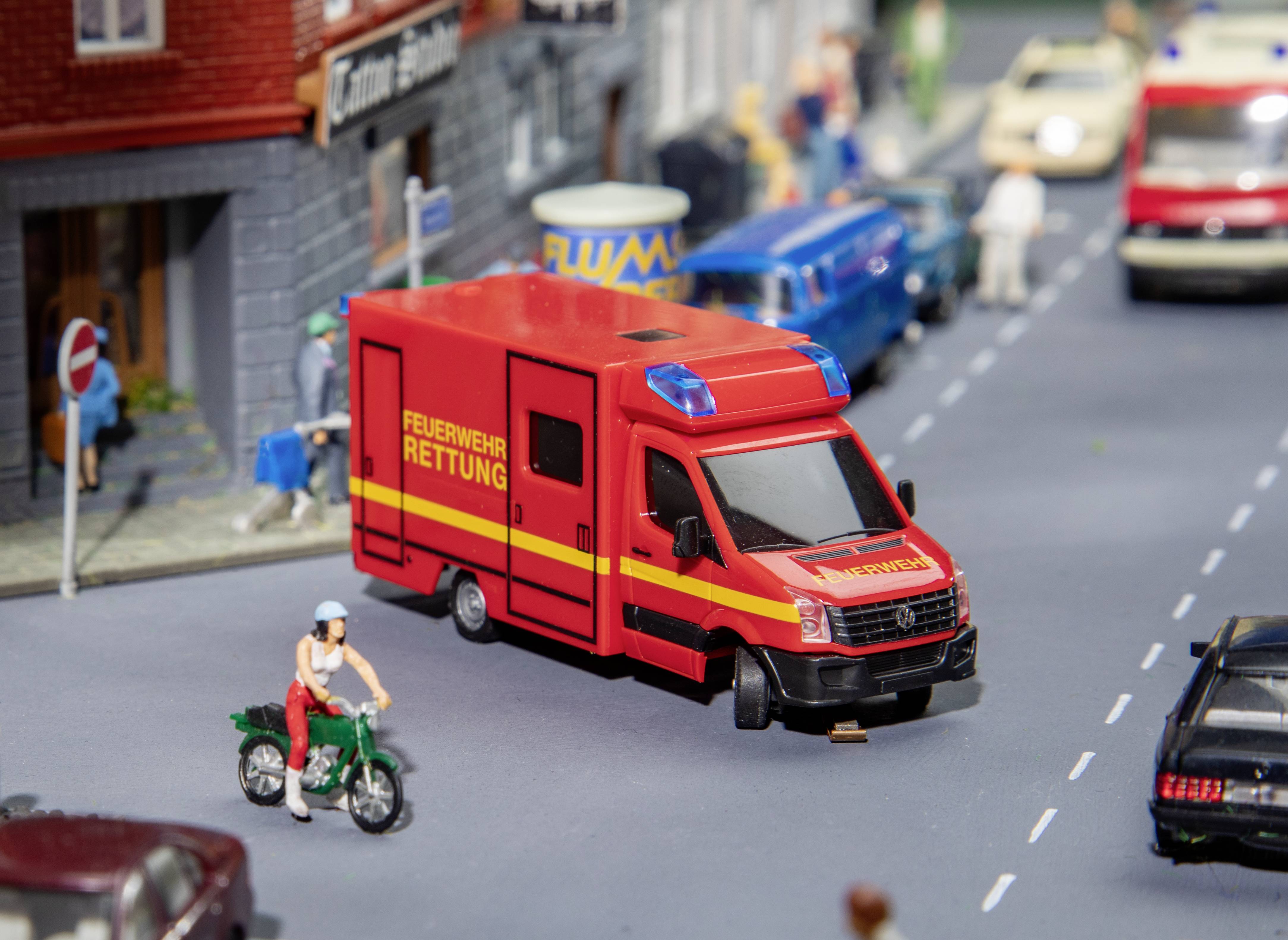 A red toy rescue vehicle with the inscription 'Fire Brigade Rescue' is positioned on a model road, surrounded by miniature vehicles.