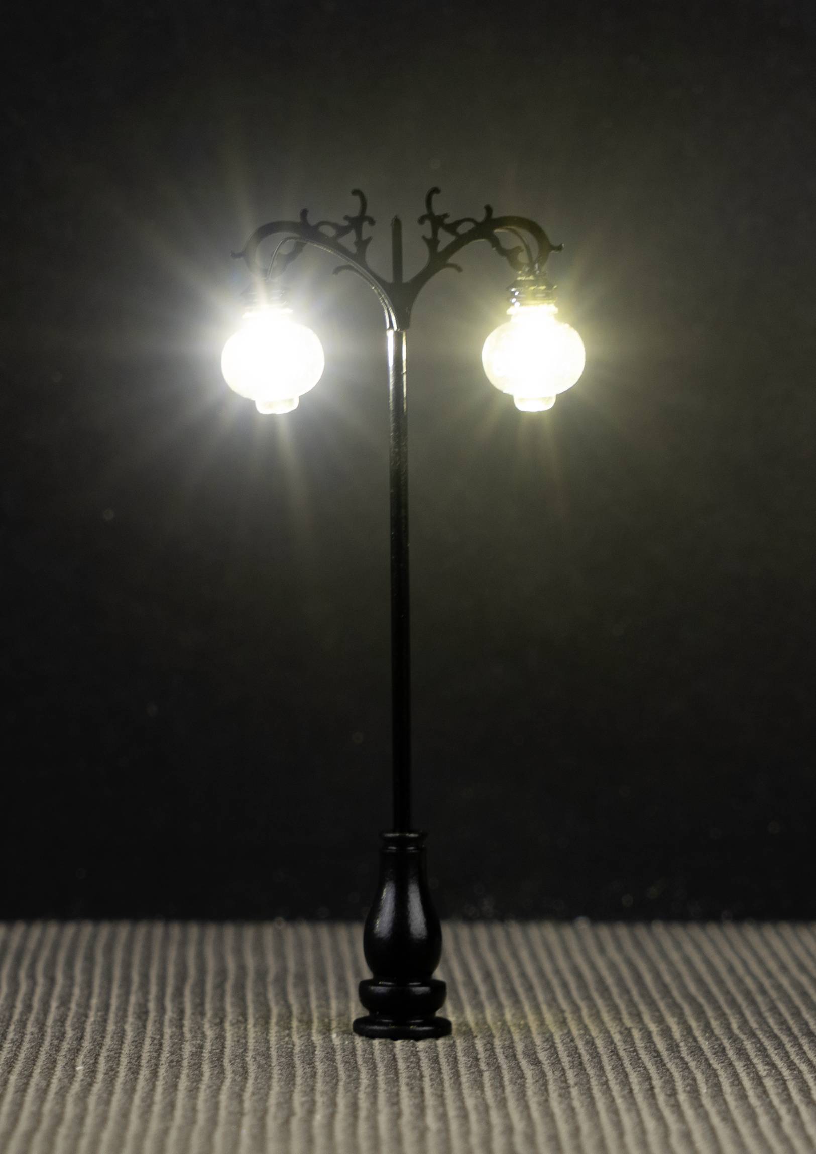 A miniature street lamp with two illuminating lights stands on a textured ground against a dark background.