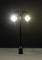 A miniature street lamp with two illuminating lights stands on a textured ground against a dark background.