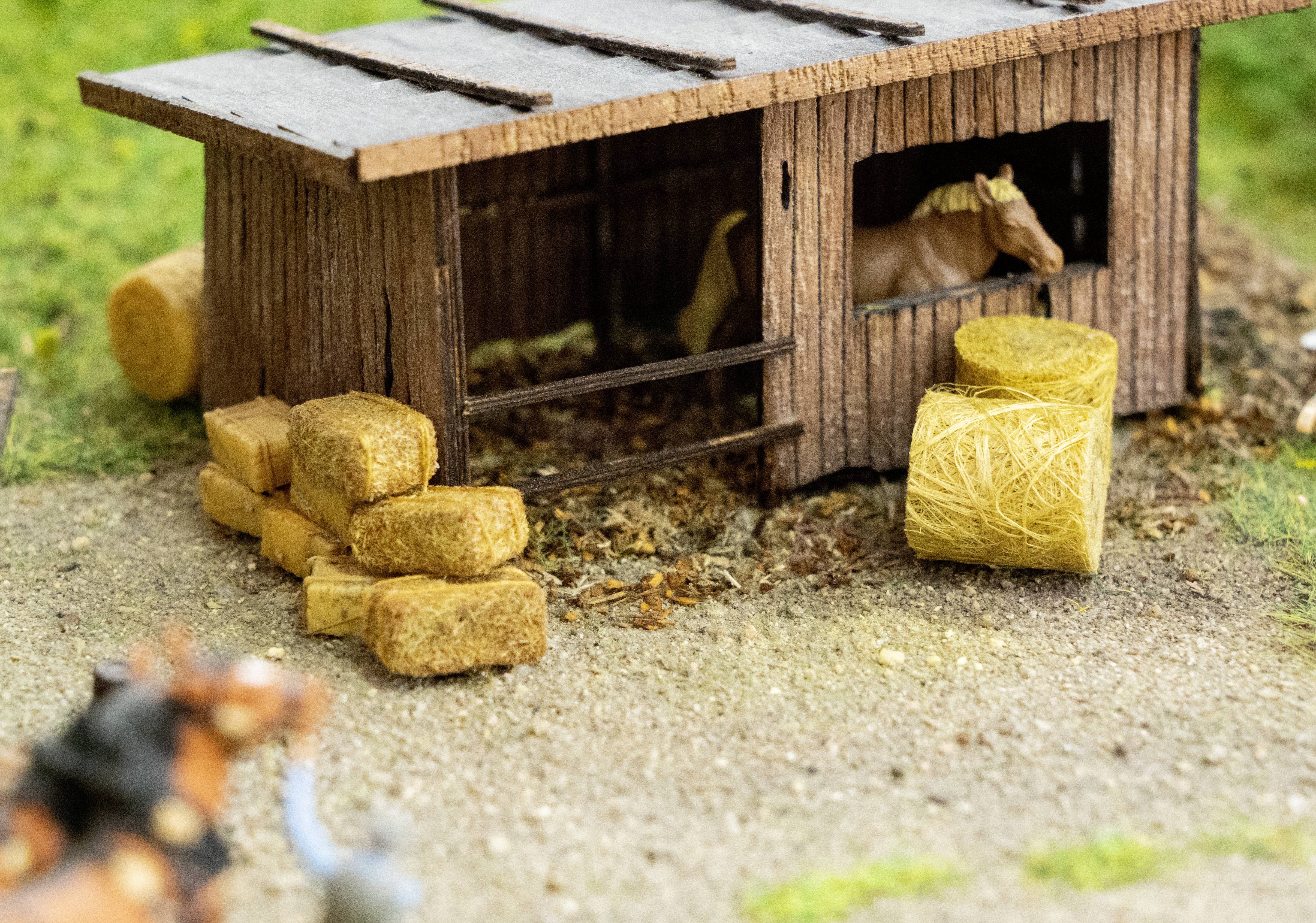 Small wooden stable with two horses. Hay bales are lying in the foreground. Surrounded by a natural, green landscape.