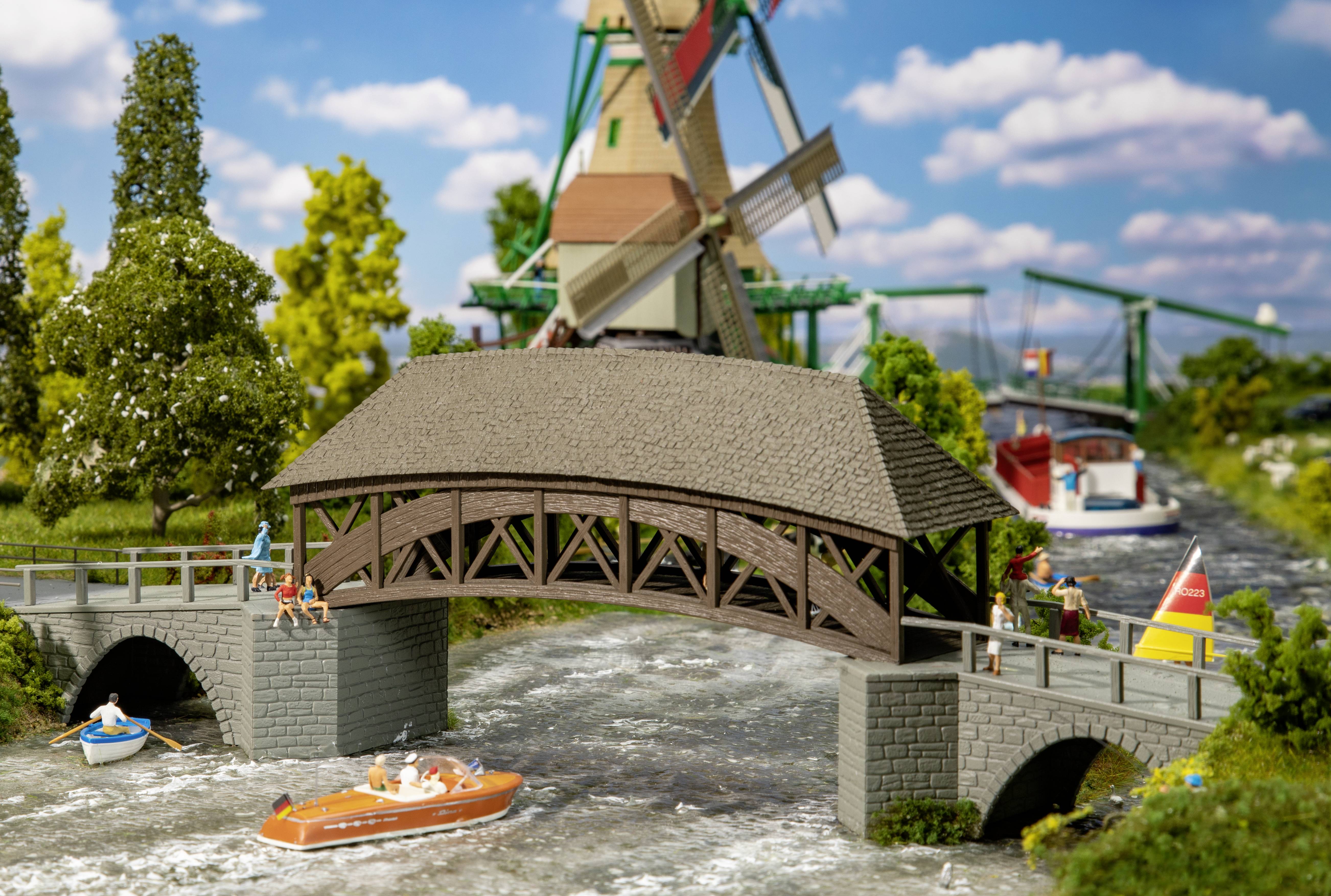 Miniature model of a picturesque river landscape with a covered wooden bridge, rowing boats, and a windmill in the background.