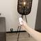 A hand holds a remote control and points it at a modern standard lamp in the room. The lamp is switched on, and the environment is minimalist.