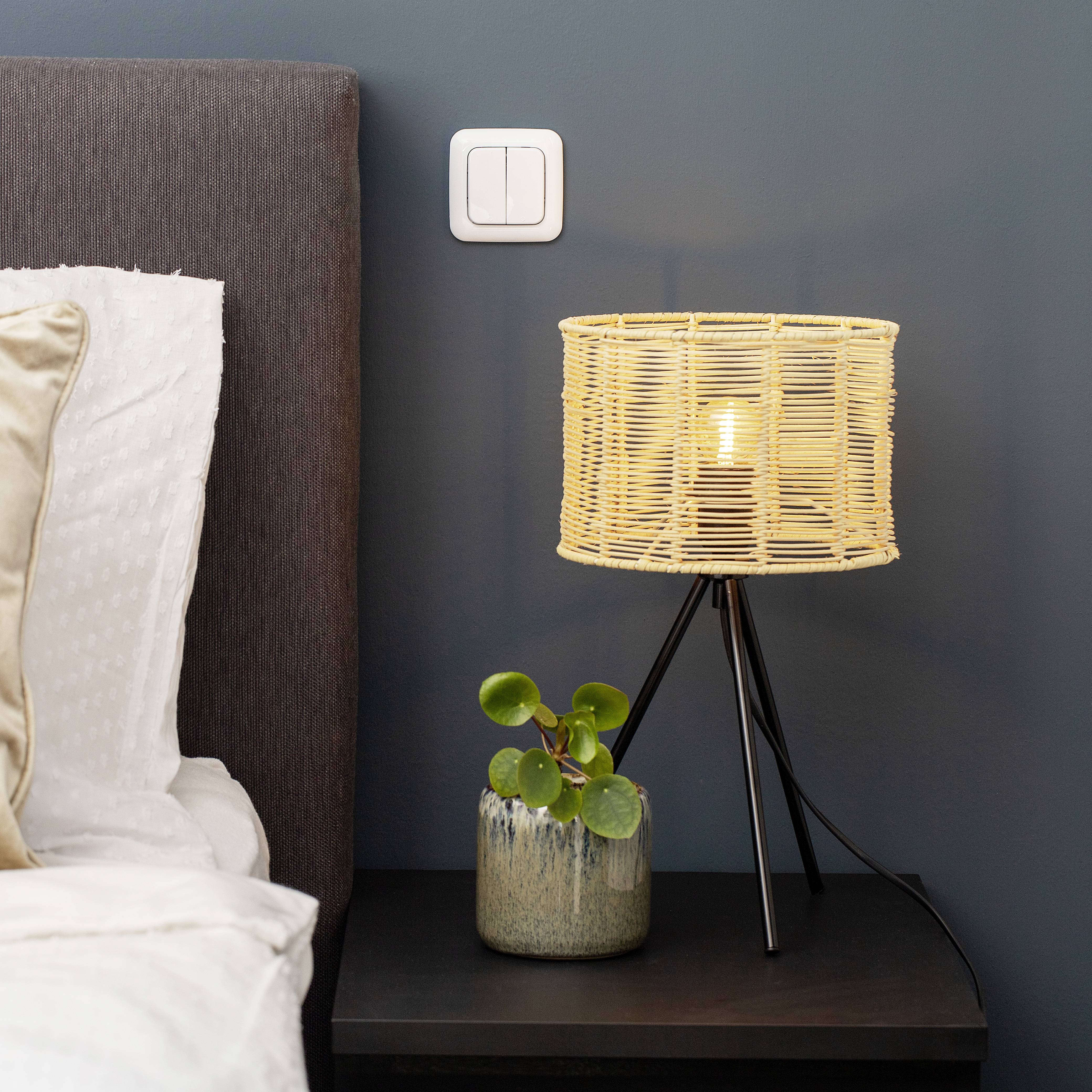 A bedside table with a lamp and a small plant stands next to a bed with grey bedding.