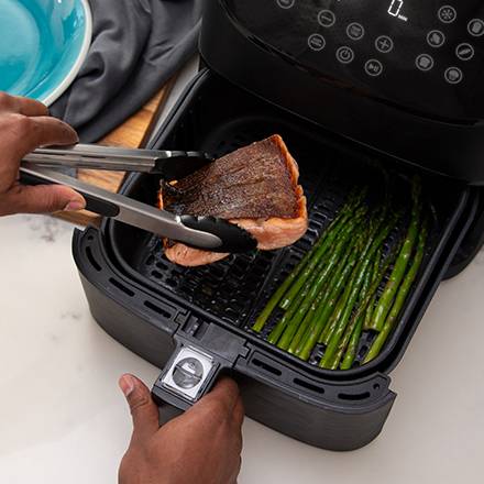 A person is placing a grilled piece of salmon and asparagus into an air fryer using tongs.