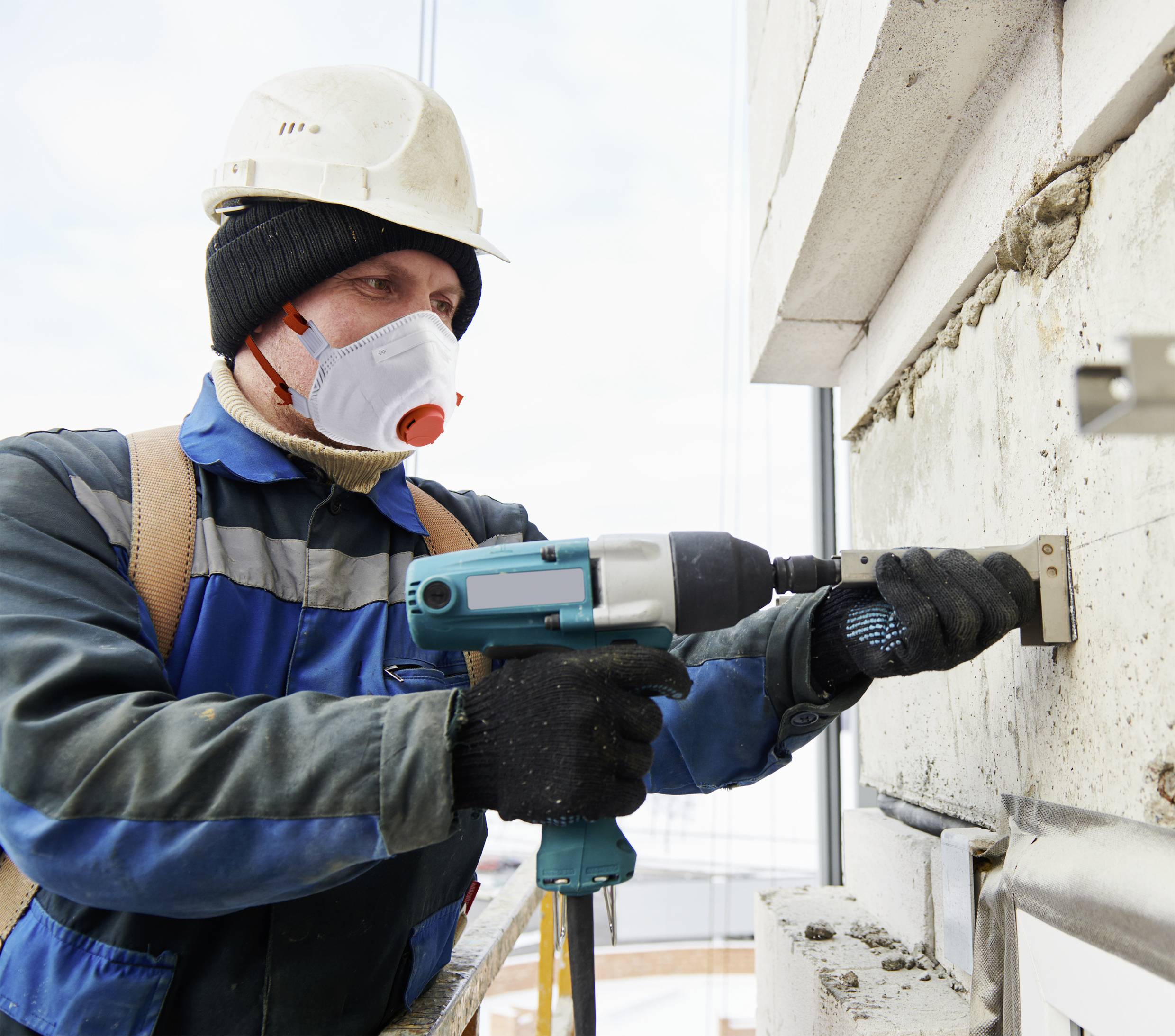 A construction worker wearing a hard hat and mask is drilling into a concrete wall with an electric tool. A scaffold can be seen behind him.