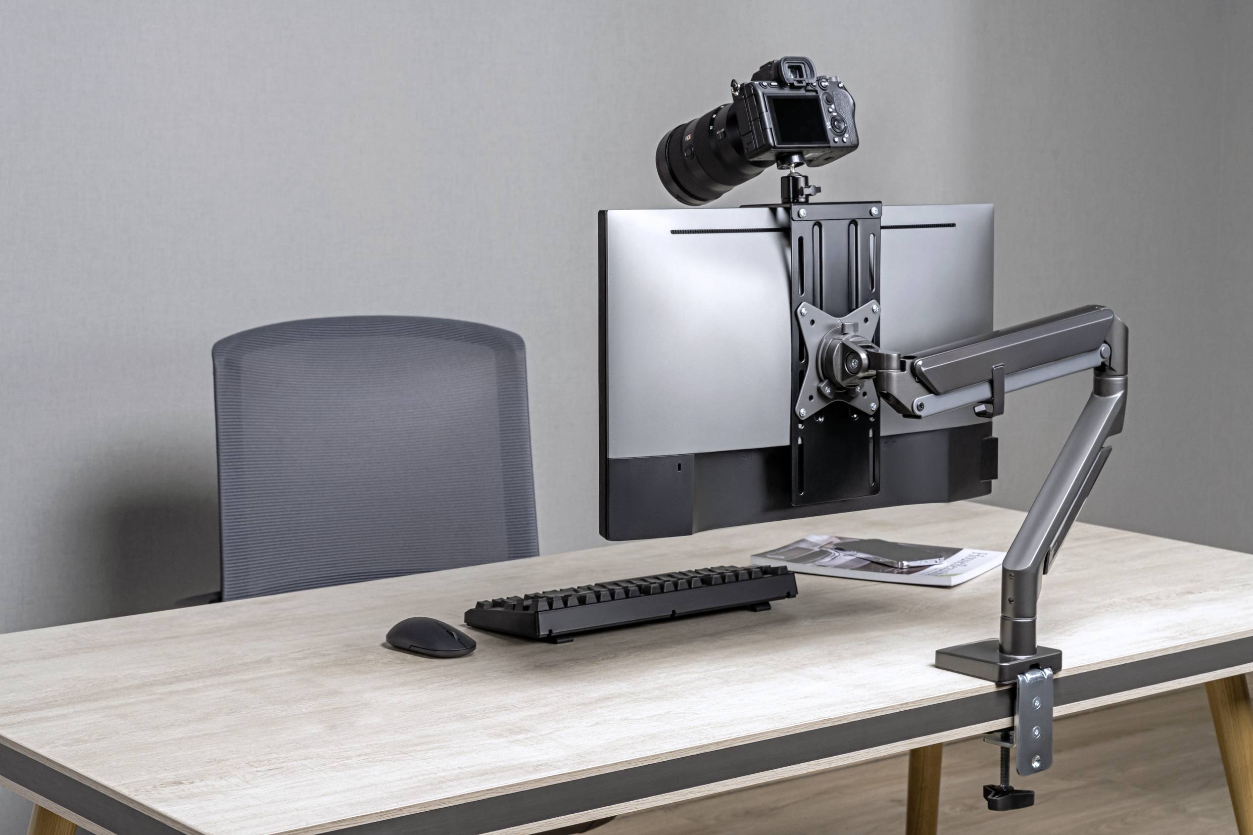 A desk with an office chair, on the table there is a monitor mount with a camera, a keyboard, a mouse and some documents.
