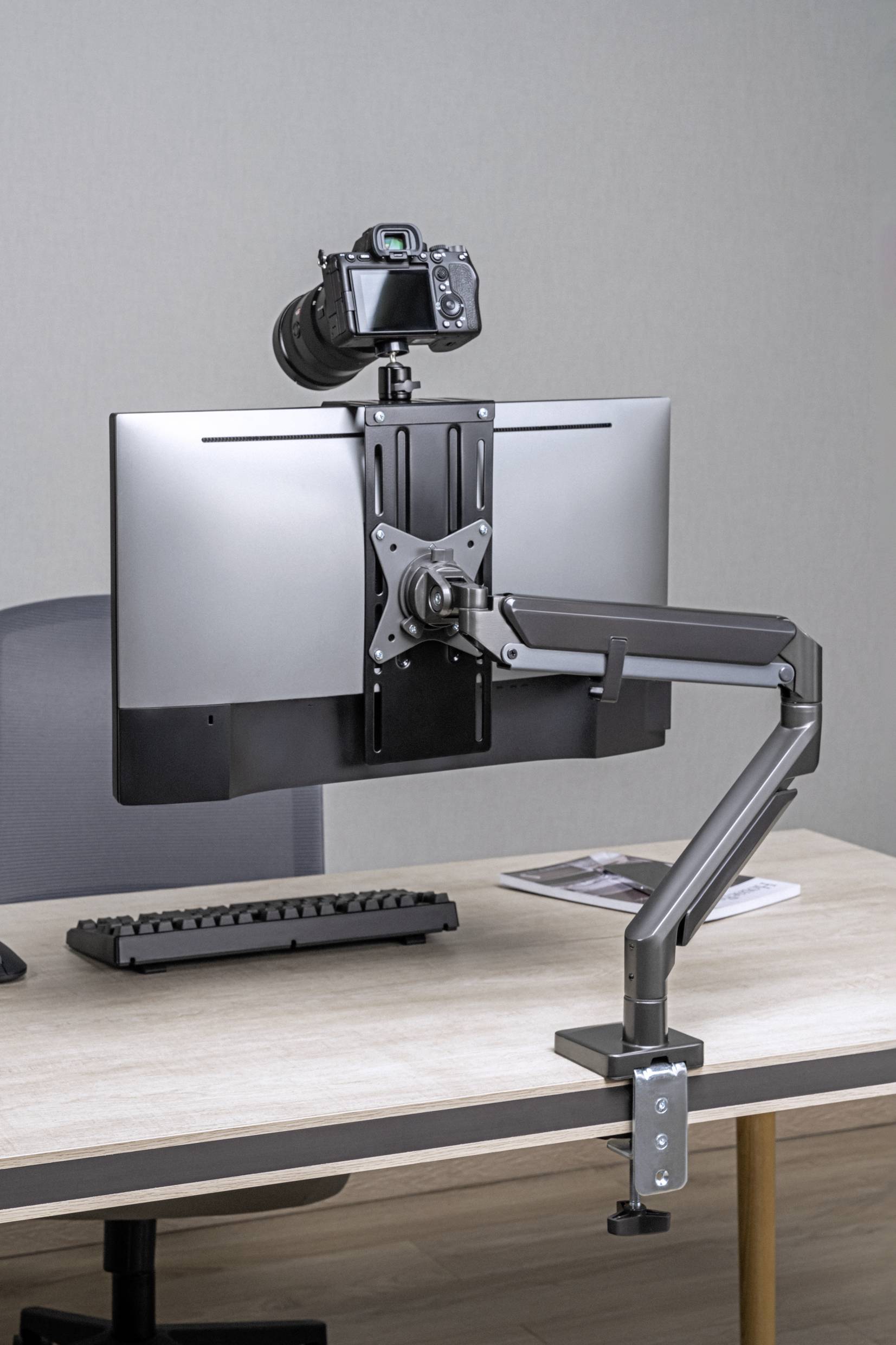 A desk with a mounted monitor arm supporting a screen. A camera is directed towards the monitor to capture contents.