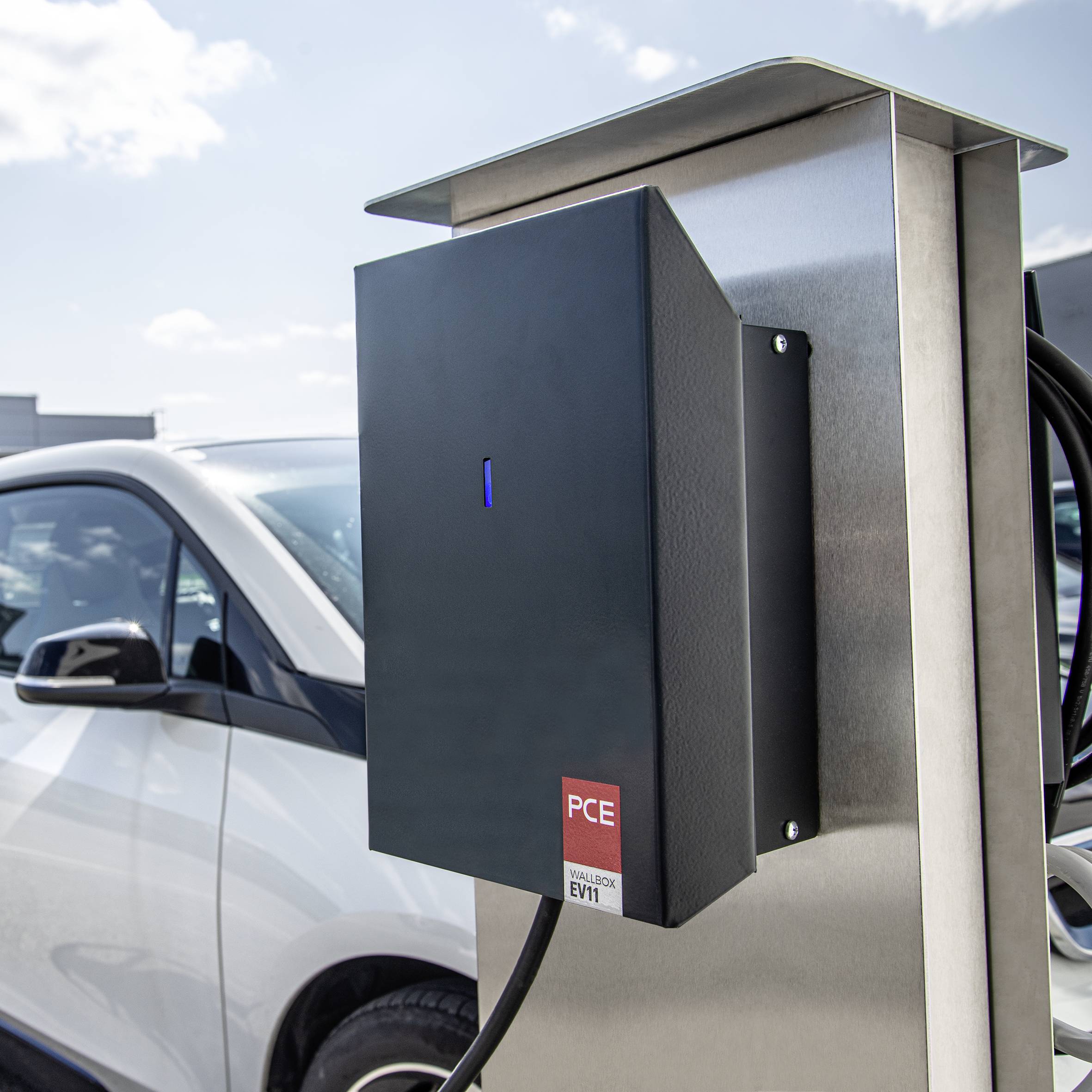 An electric car is being charged at a charging station in a car park. Further vehicles and buildings are visible in the background.