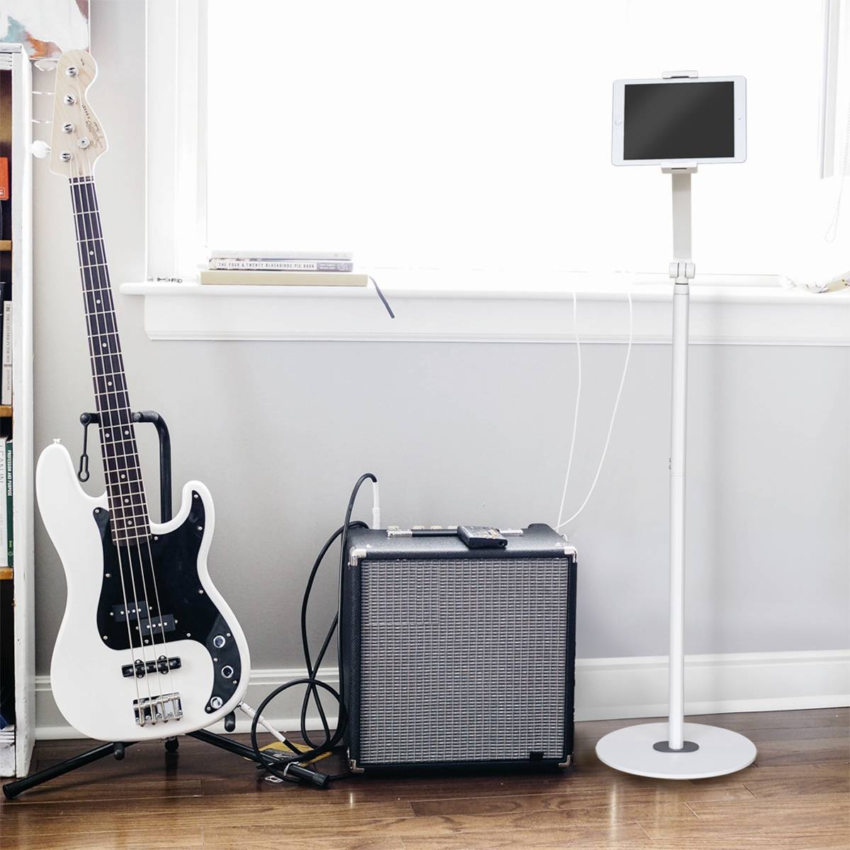 Electric bass guitar, amplifier and tablet on a stand are positioned side by side on a wooden floor in front of a window.