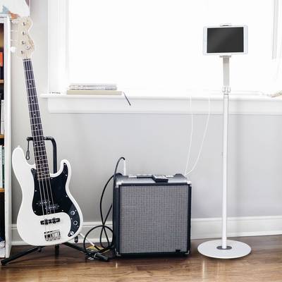 Electric bass guitar, amplifier and tablet on a stand are positioned side by side on a wooden floor in front of a window.