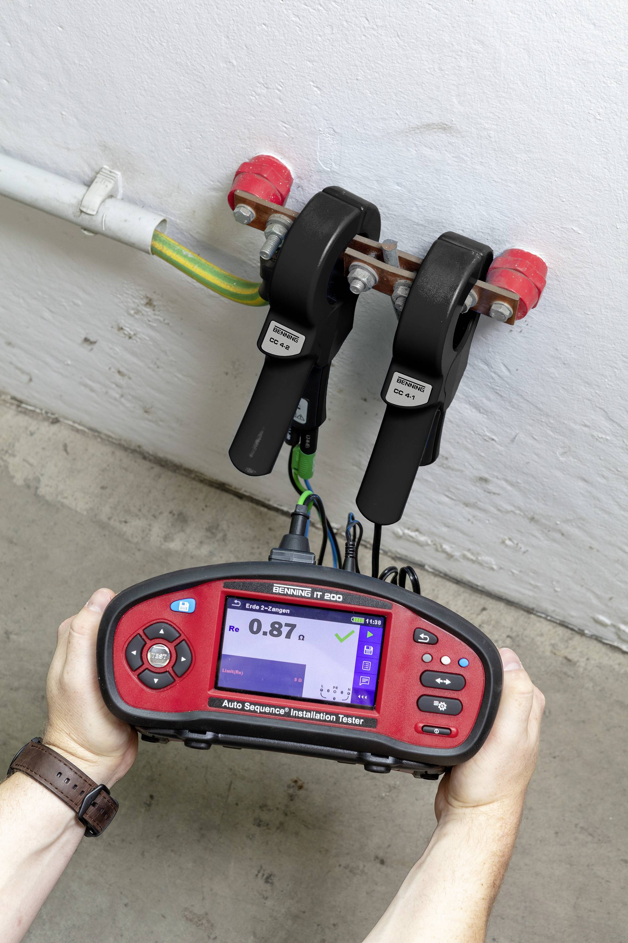 A person is holding an electrical testing device, measuring two cables fixed to a wall. The display shows '0.87'.
